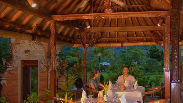 Un área de comedor al aire libre con una estructura de madera cubierta, mesas dispuestas con manteles blancos y vistas a exuberante follaje de fondo.