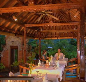 An outdoor dining area with a covered wooden structure, tables set with white tablecloths, and a view of lush greenery in the background. - Puri Bagus Manggis (Manggis)