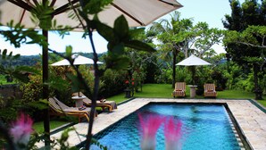 Una piscina con sillas de descanso y sombrillas, rodeada de exuberante vegetación.