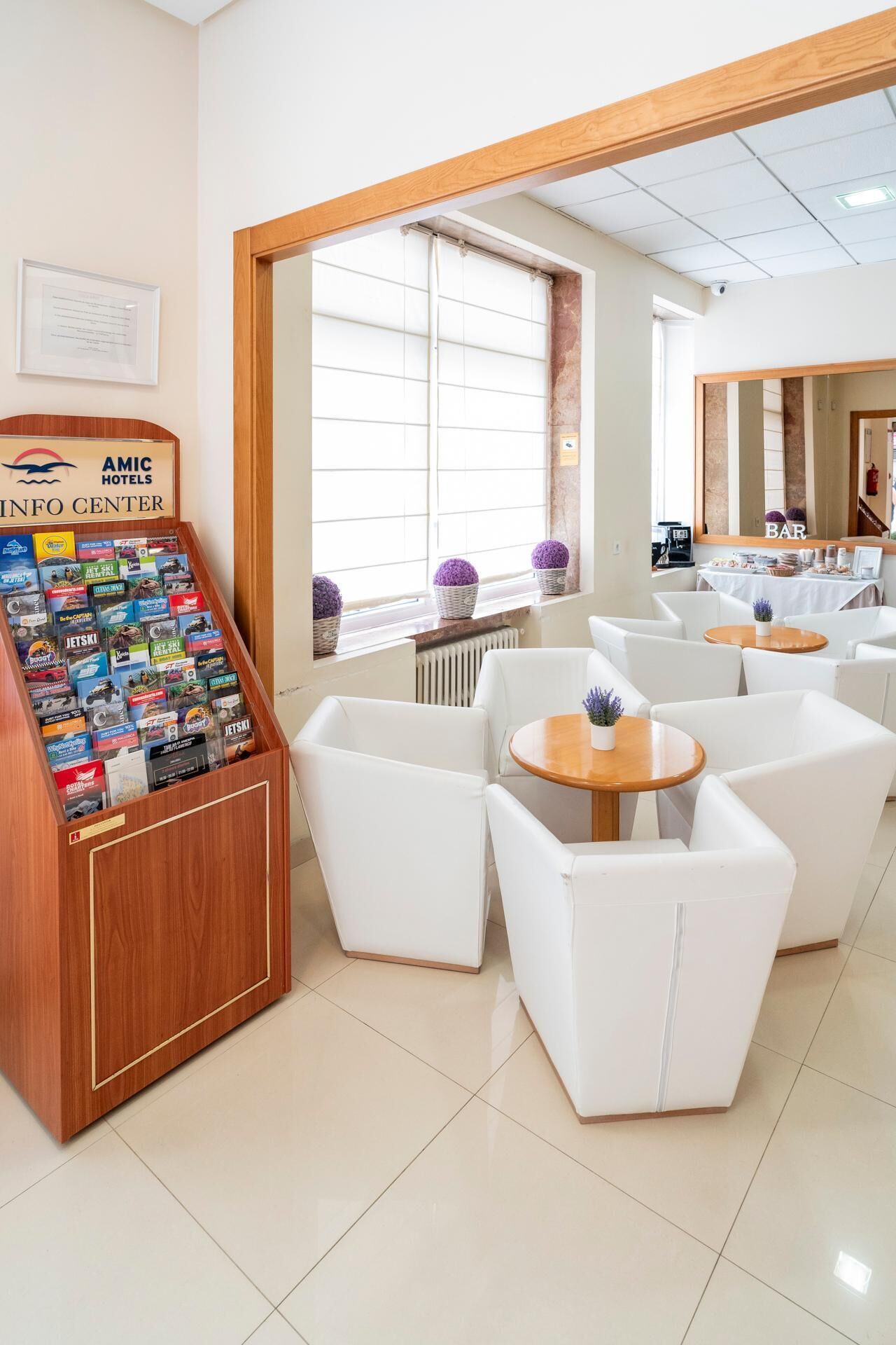Et hotellobby med en receptionsdisk, et opholdsområde med hvide stole og borde, samt et stort vindue med persienner.