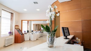 A reception area with a white reception desk, a glass vase with white orchids, a wooden wall, and a bar area in the background. - Hotel Amic Colón (Palma de Mallorca)
