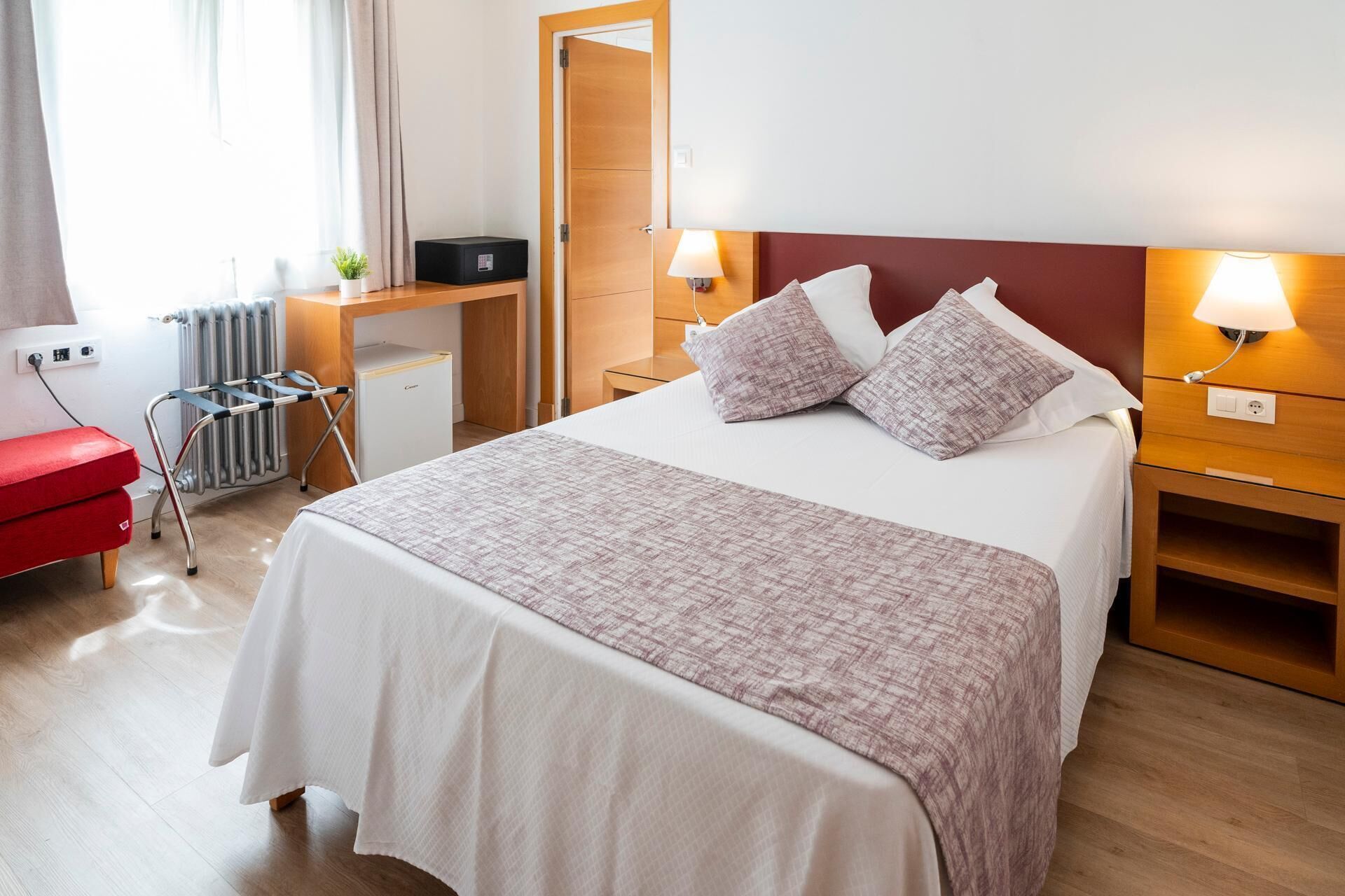 Habitación de hotel con cama, mesitas de noche, escritorio, radiador, una silla roja y una planta pequeña.