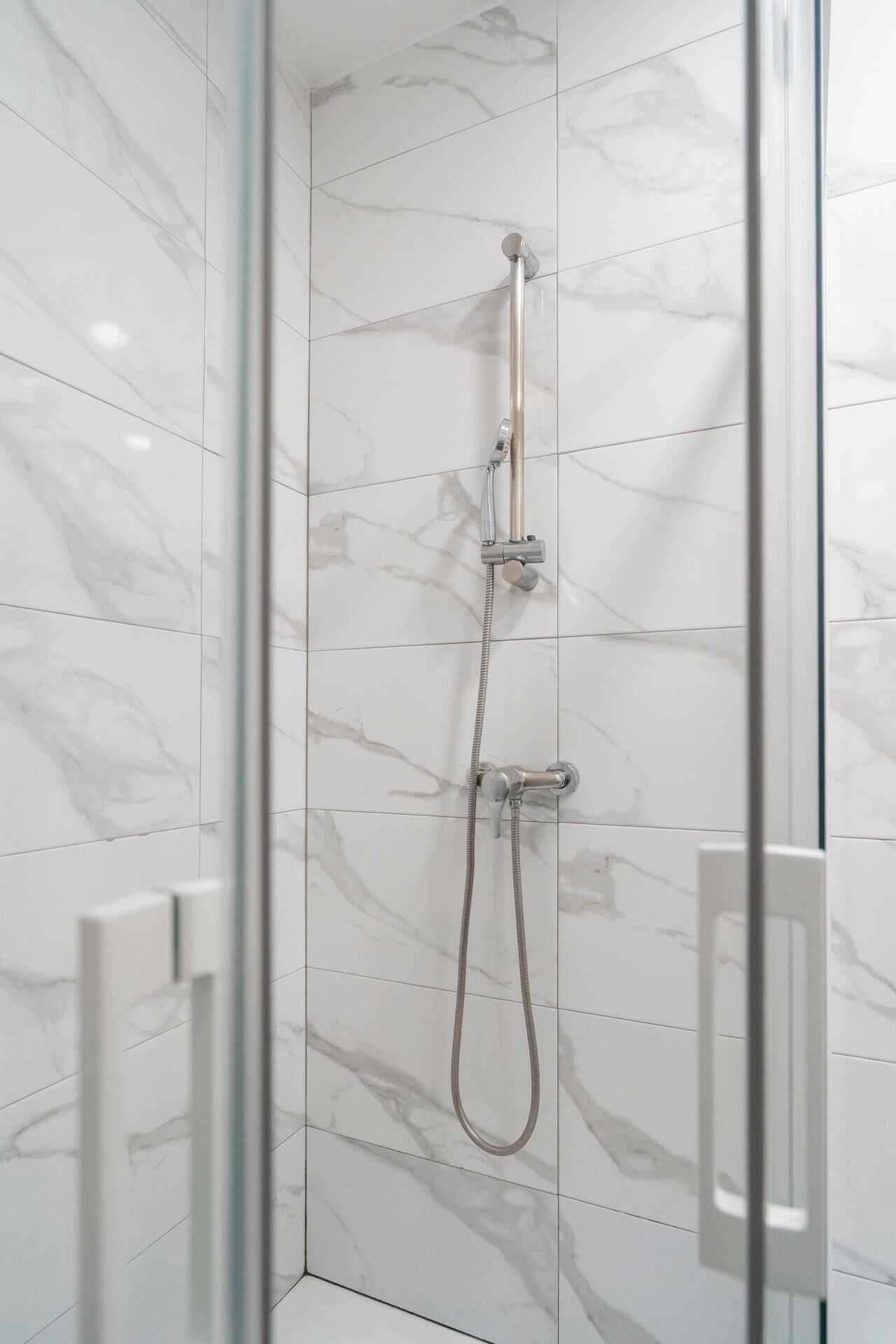 Ducha envuelta en vidrio con cabezal de ducha manual y cabezal de ducha fijo, frente a una pared de azulejos de mármol blanco.