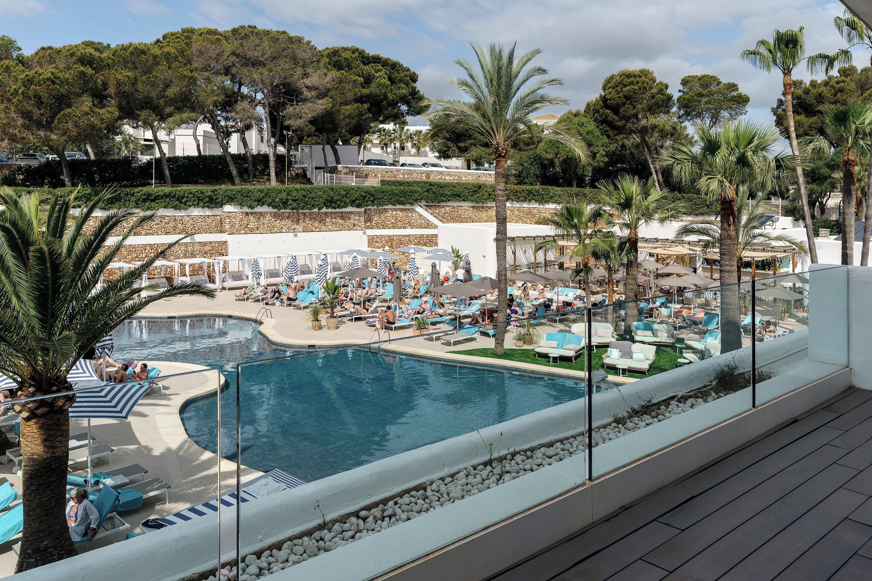 A poolside area with lounge chairs and umbrellas, surrounded by palm trees and a stone wall.