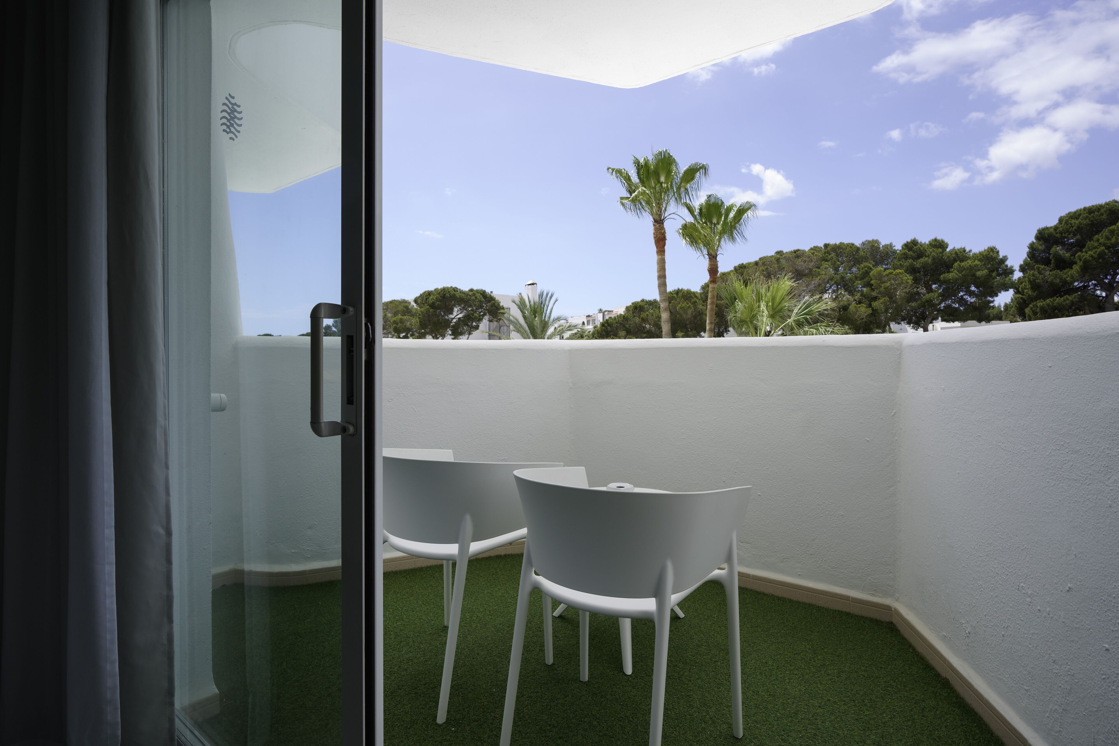 A glass door leading to a balcony with two white chairs and artificial grass.