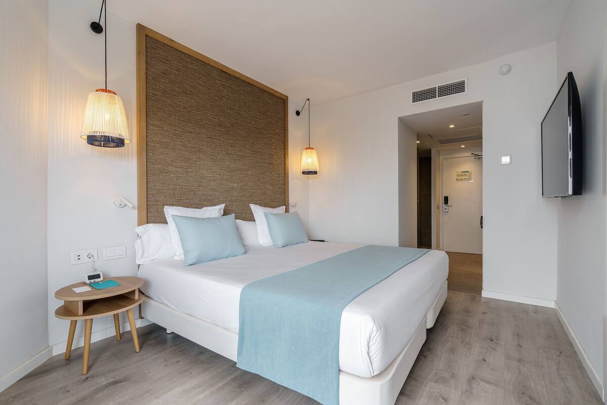 Habitación de hotel ordenada con una cama grande, lámparas de noche, una mesita de madera pequeña y un televisor de pantalla plana montado en la pared.