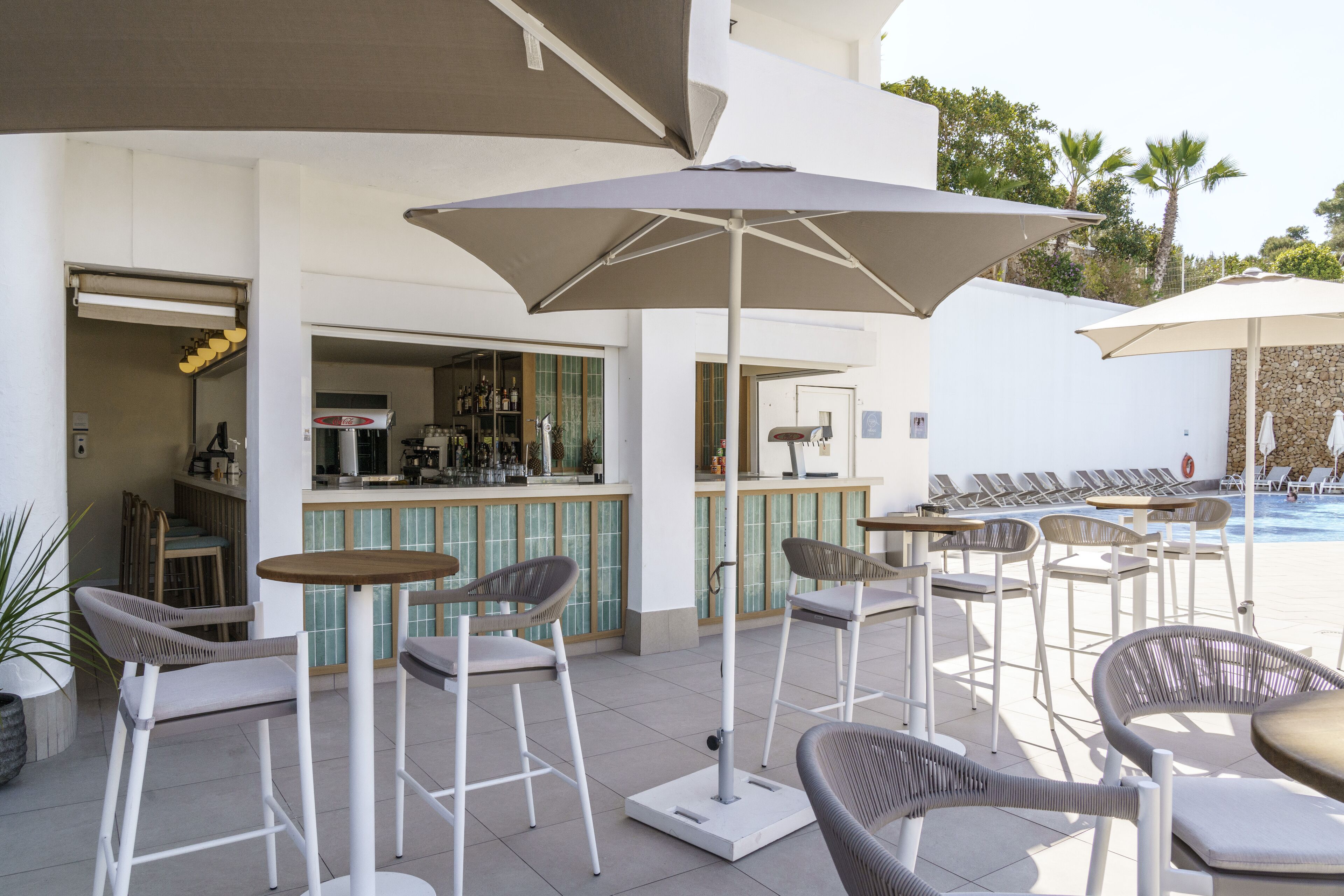 An outdoor bar area with seating and umbrellas, a pool in the background, and a bar with a tiled backsplash.