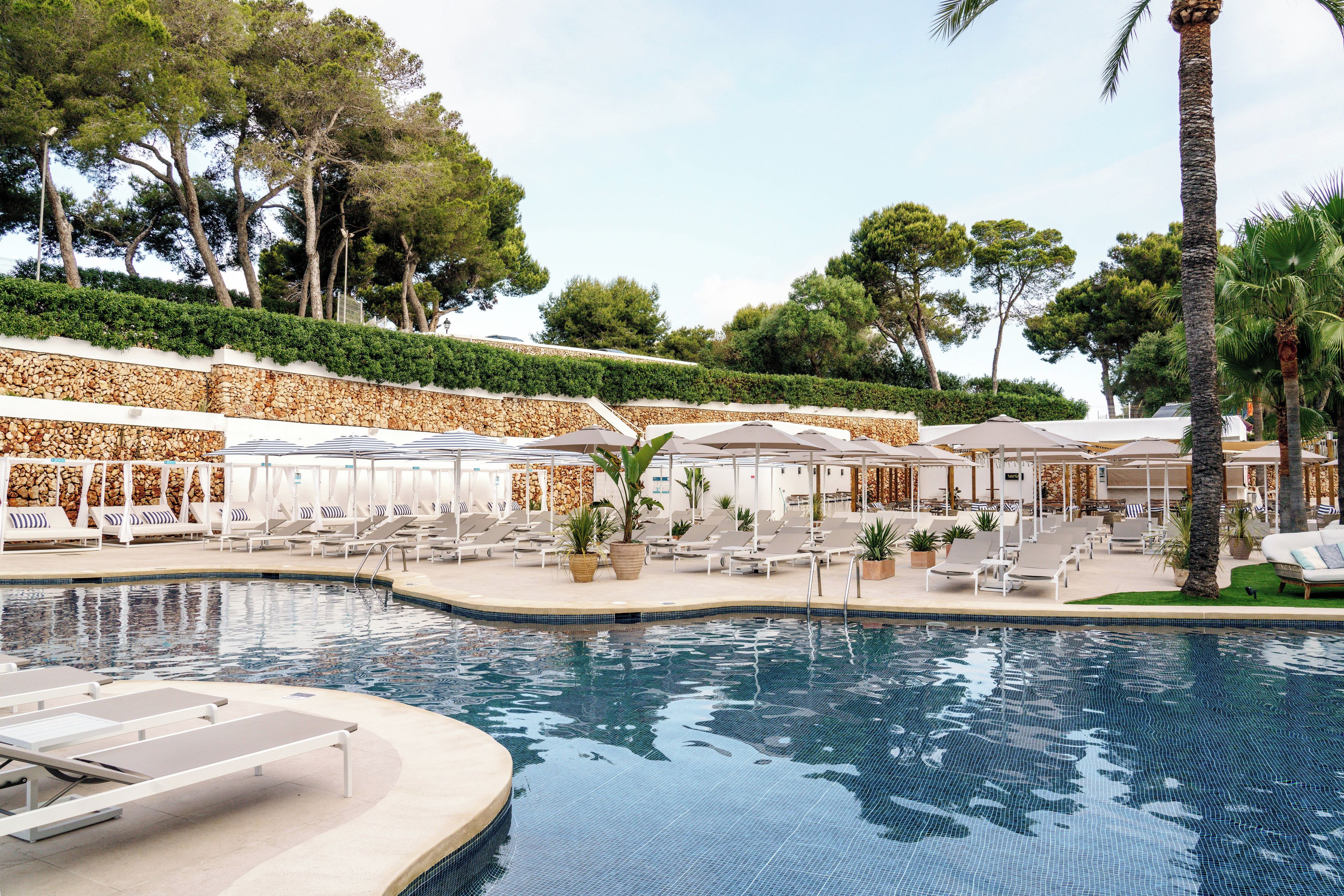 Un área de piscina con sillones de descanso y sombrillas, rodeada de árboles y un muro de piedra.