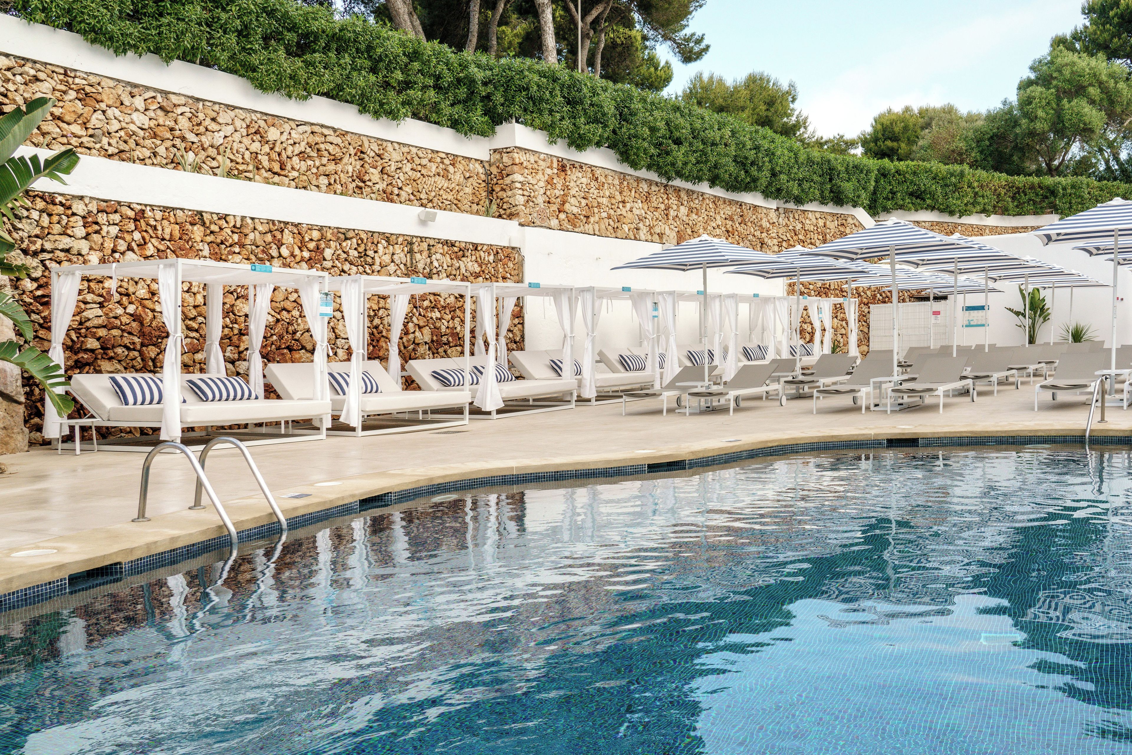 Zona junto a la piscina con sillas de descanso y sombrillas, una pared de piedra y una piscina cristalina.