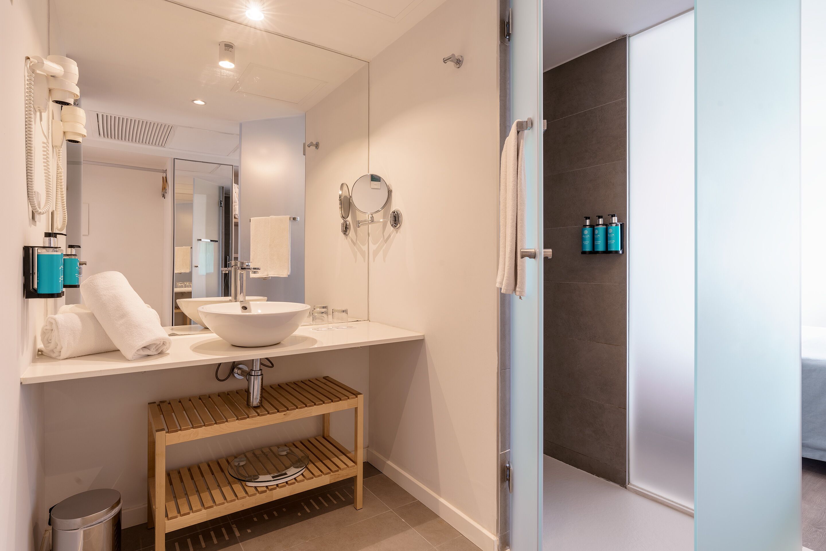 Ein modernes Badezimmer mit einem weißen Waschbecken, Spiegel und einem Holzbodenregal. An der Wand befinden sich Pflegeprodukte und ein Handtuch auf der Ablagefläche.