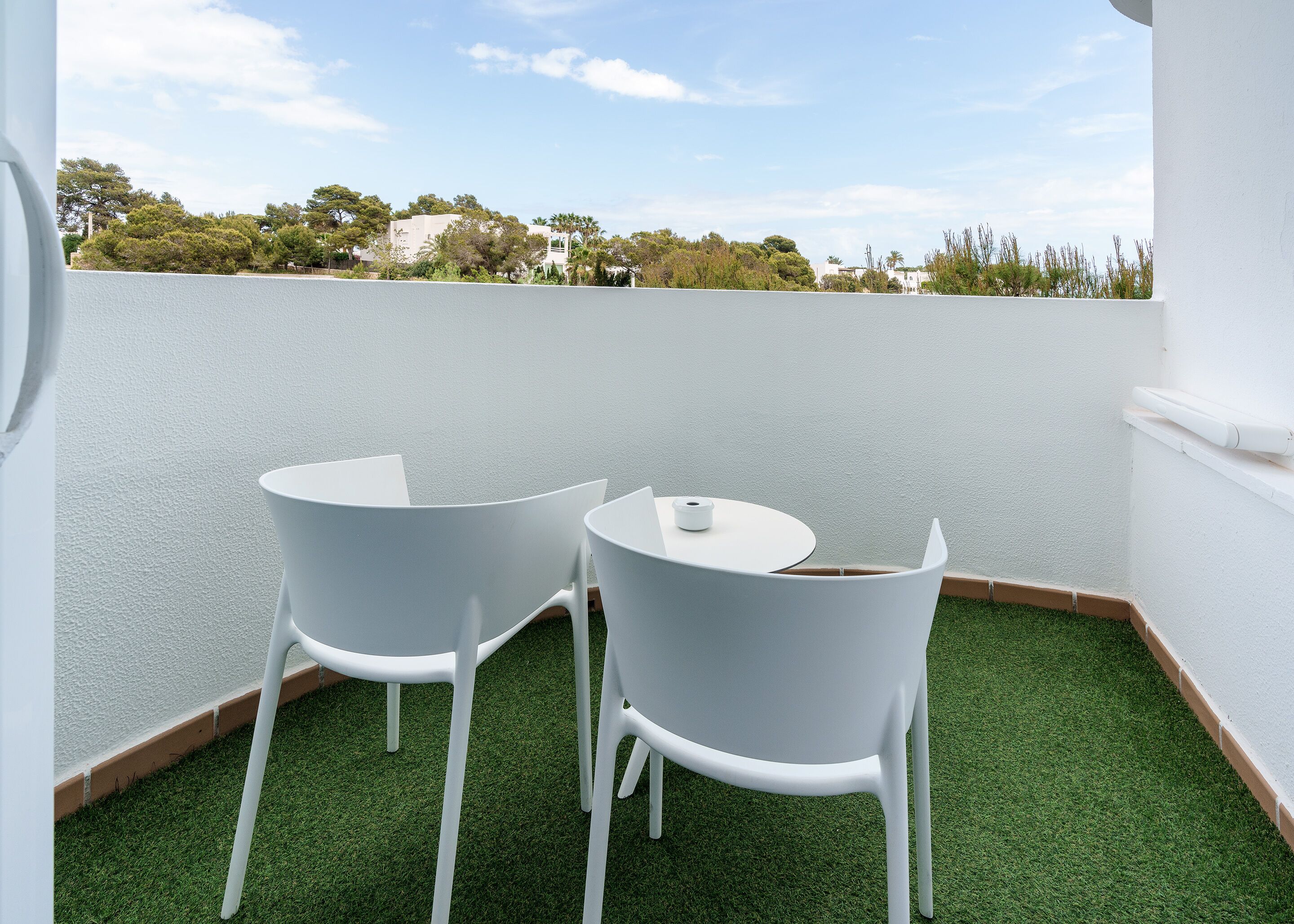 A small outdoor seating area with two white chairs and a table, surrounded by a white wall and a view of trees and buildings in the background.