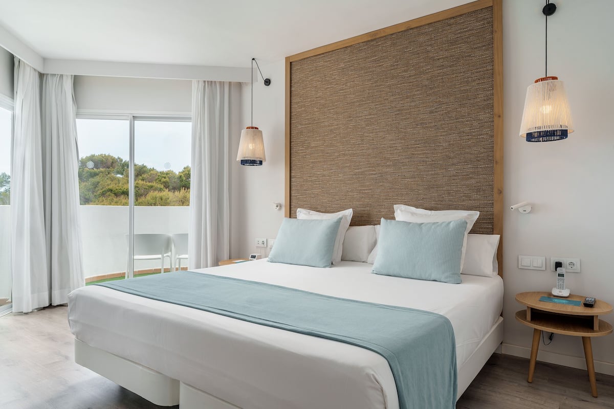 Habitación de hotel moderna con una cama grande, un cabecero de madera, una mesita de noche pequeña con un teléfono y vistas a zonas verdes.