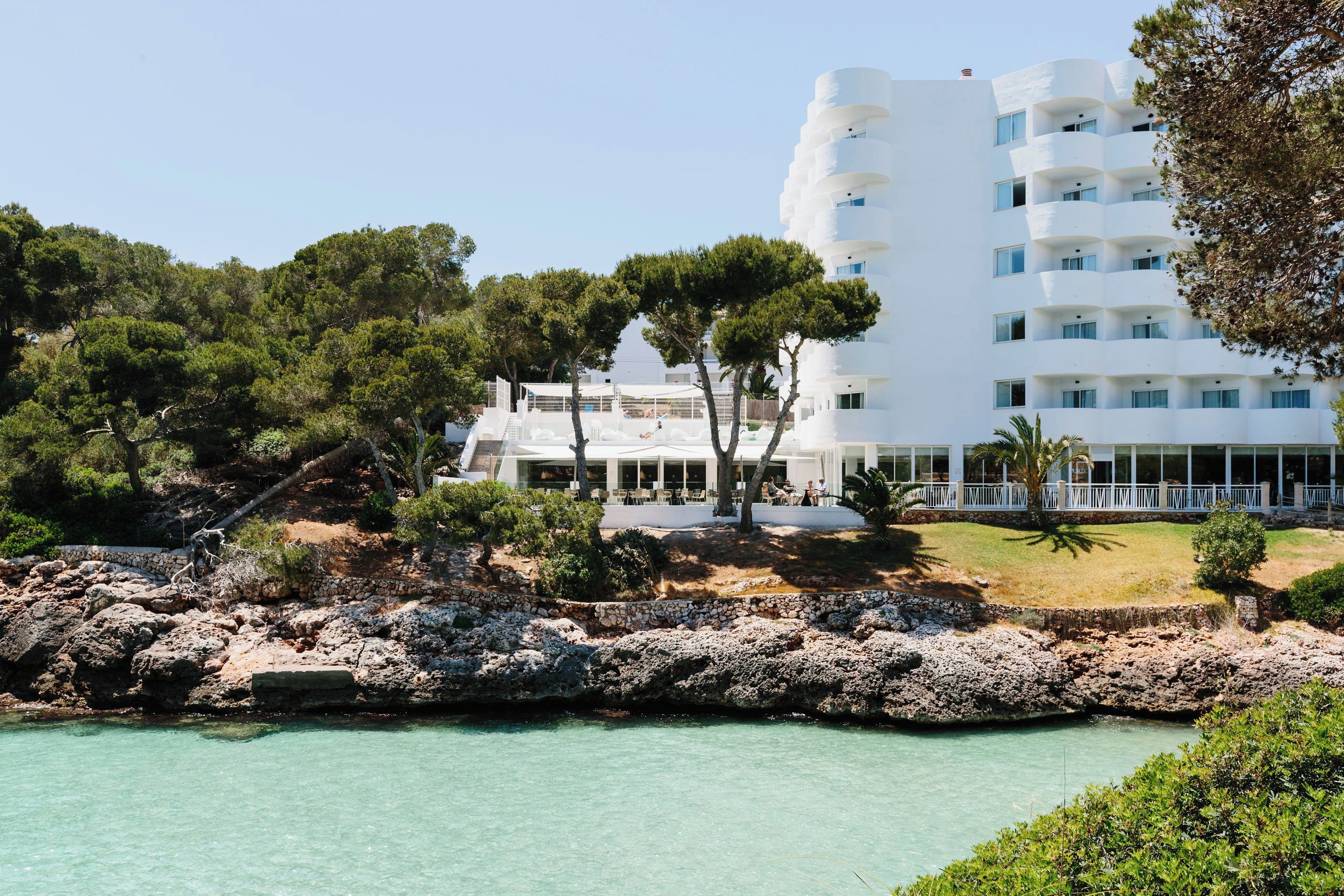 A coastal hotel with a white facade, multiple balconies, and a rocky shoreline.