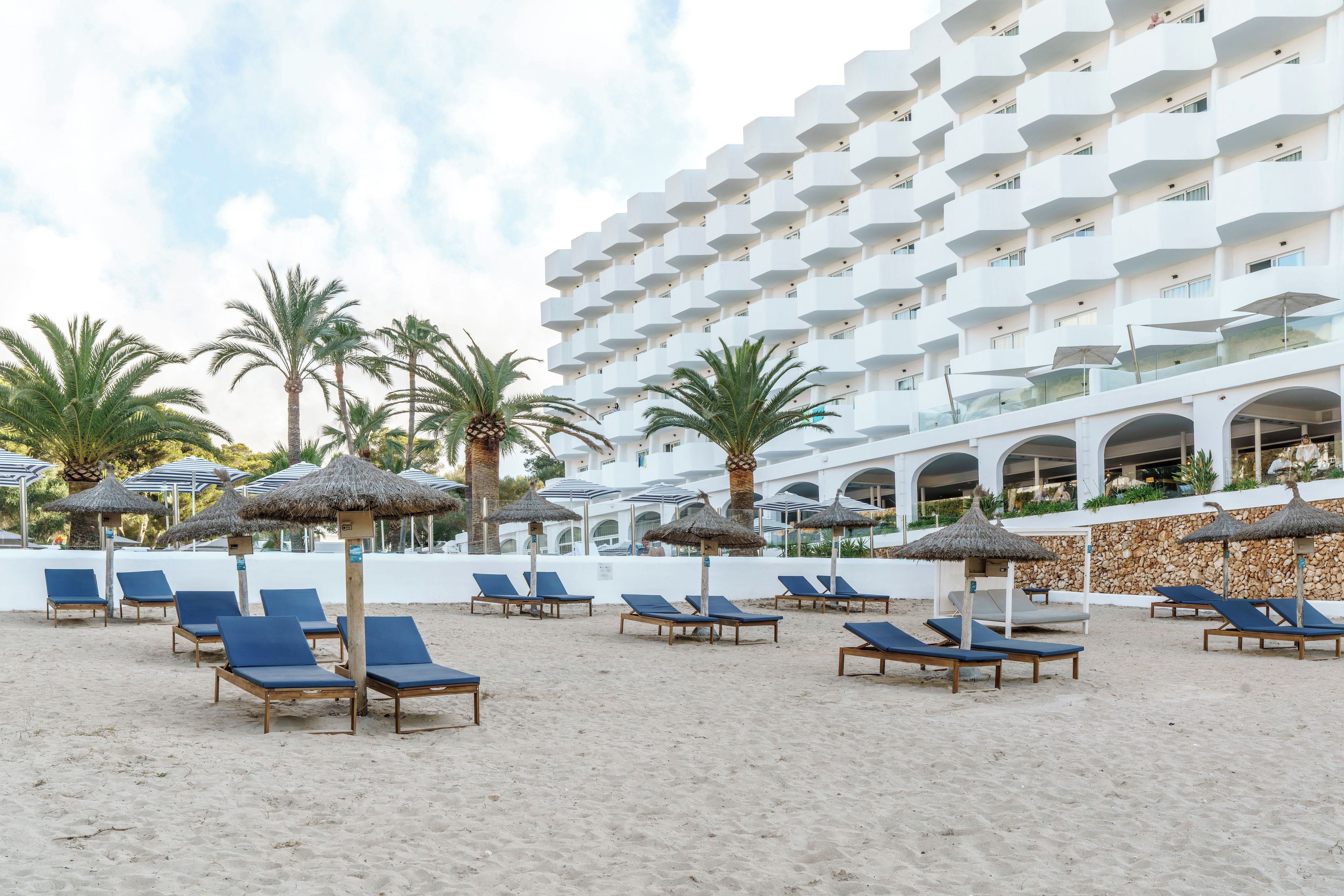 Ein Hotel direkt am Strand mit mehreren Balkonen, Liegestühlen und Reetdachschirmen.