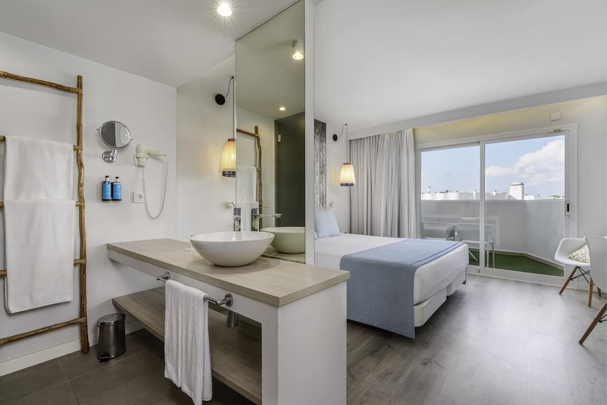 Una habitación de hotel moderna con una cama grande, un baño con lavamanos y espejo, y un balcón con vistas.