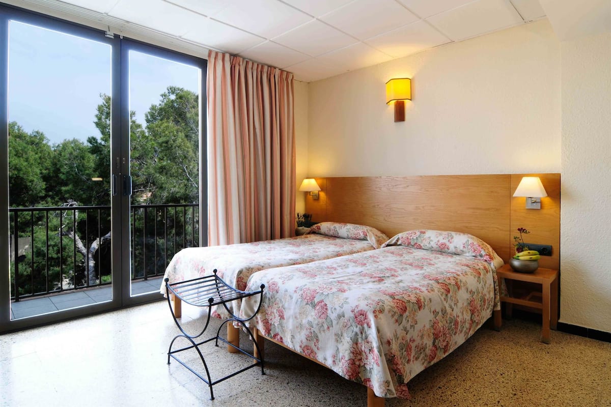 A hotel room with two beds, a balcony, and a view of trees.