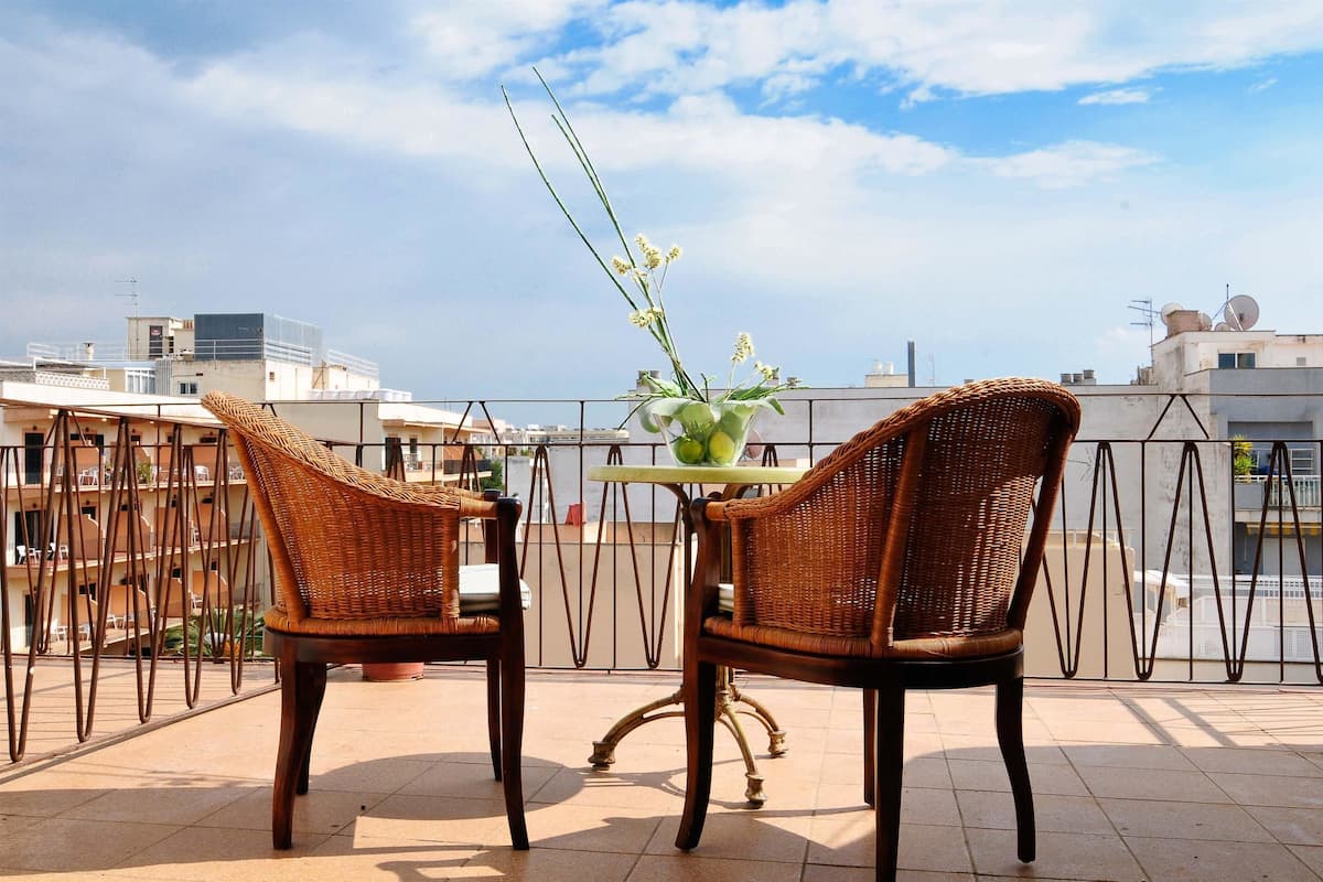 A balcony with wicker chairs and a table, a vase with flowers, and a view of buildings in the background.