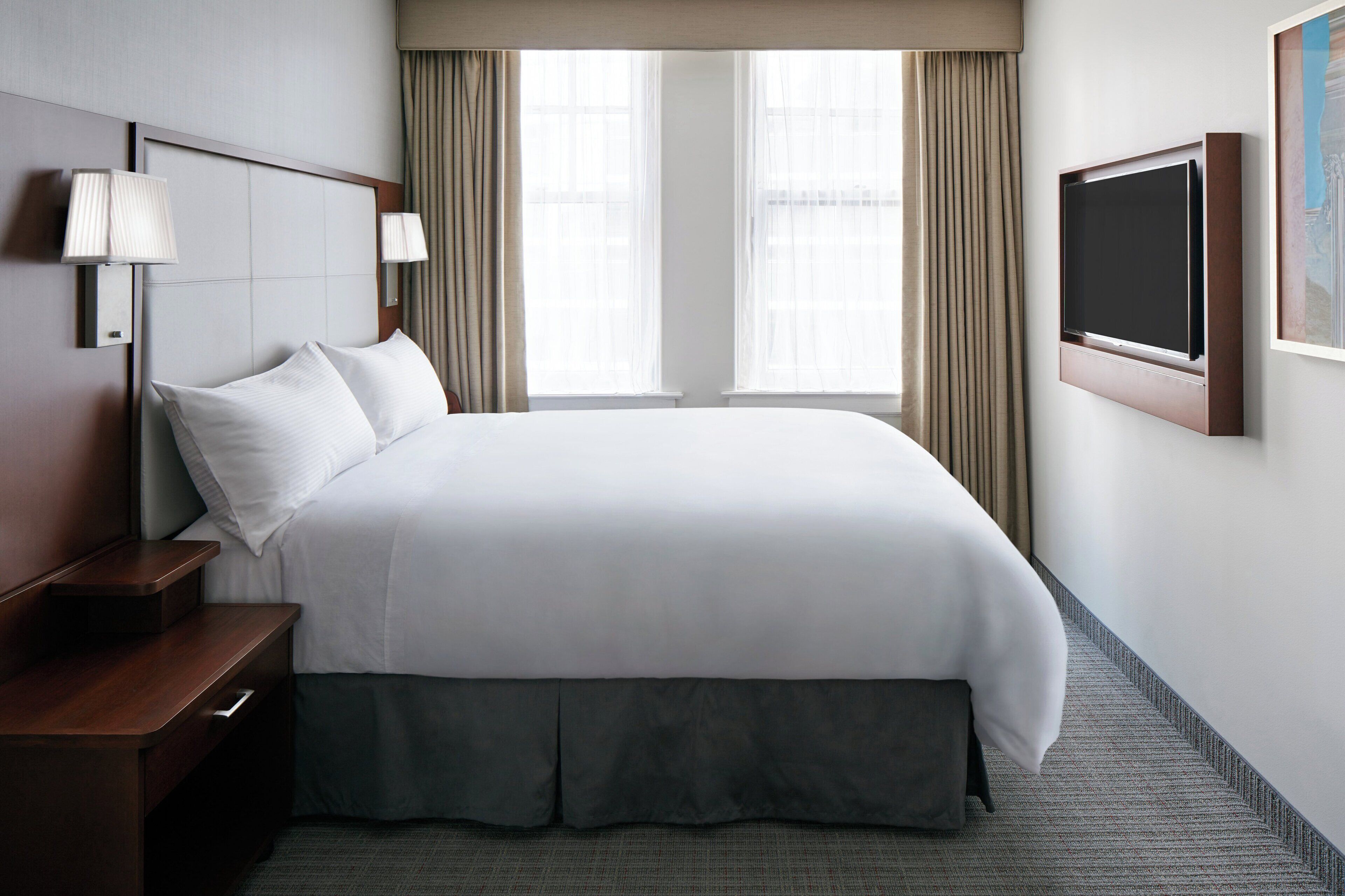 Een hotelkamer met een groot bed, een nachtkastje, een aan de muur bevestigde televisie en een raam met gordijnen.