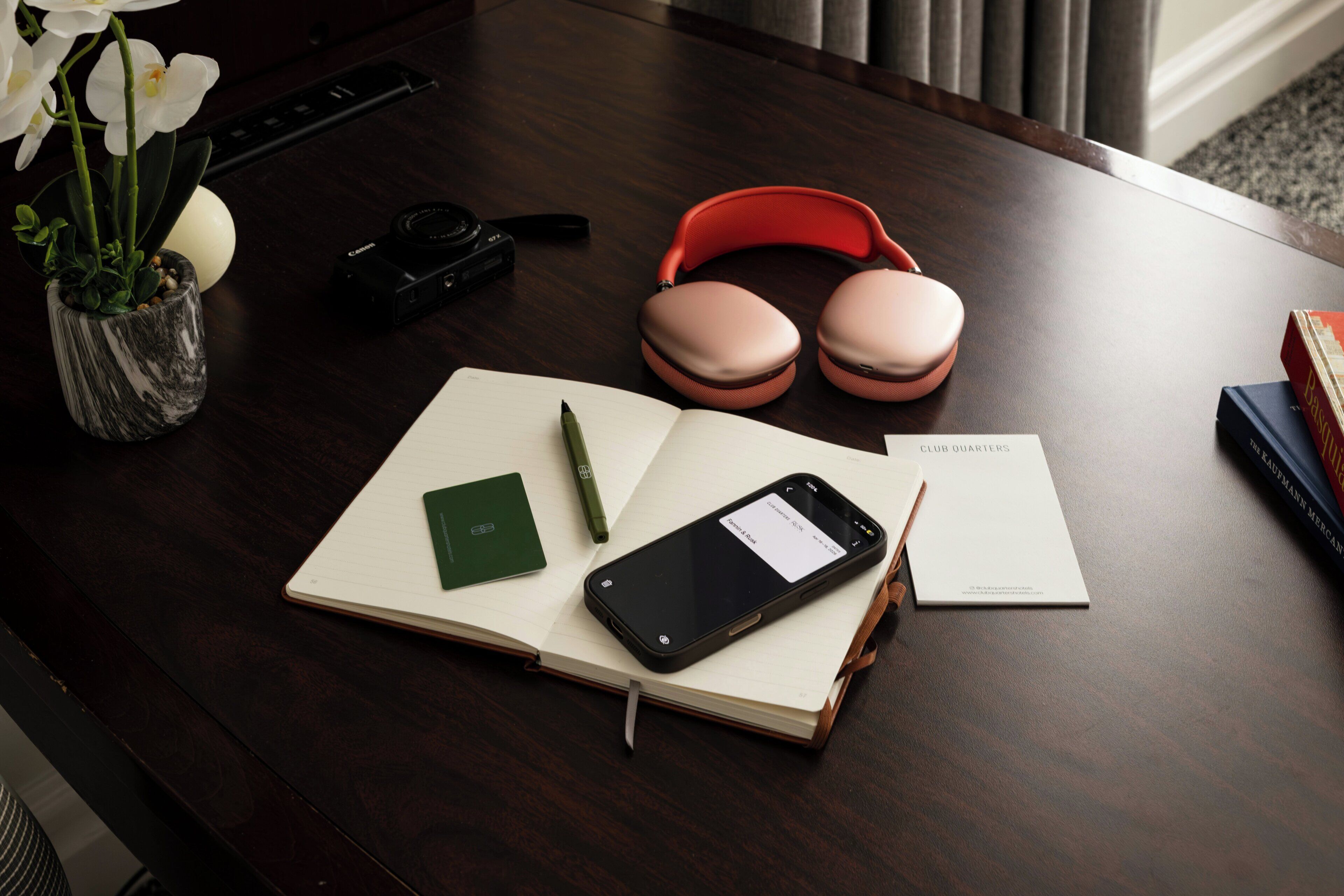 A wooden desk with a notebook, a smartphone, a pen, a pair of headphones, a camera, and a vase with flowers.