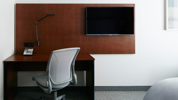A hotel room with a wooden desk, a modern chair, a flat-screen TV mounted on the wall, and a telephone on the desk.