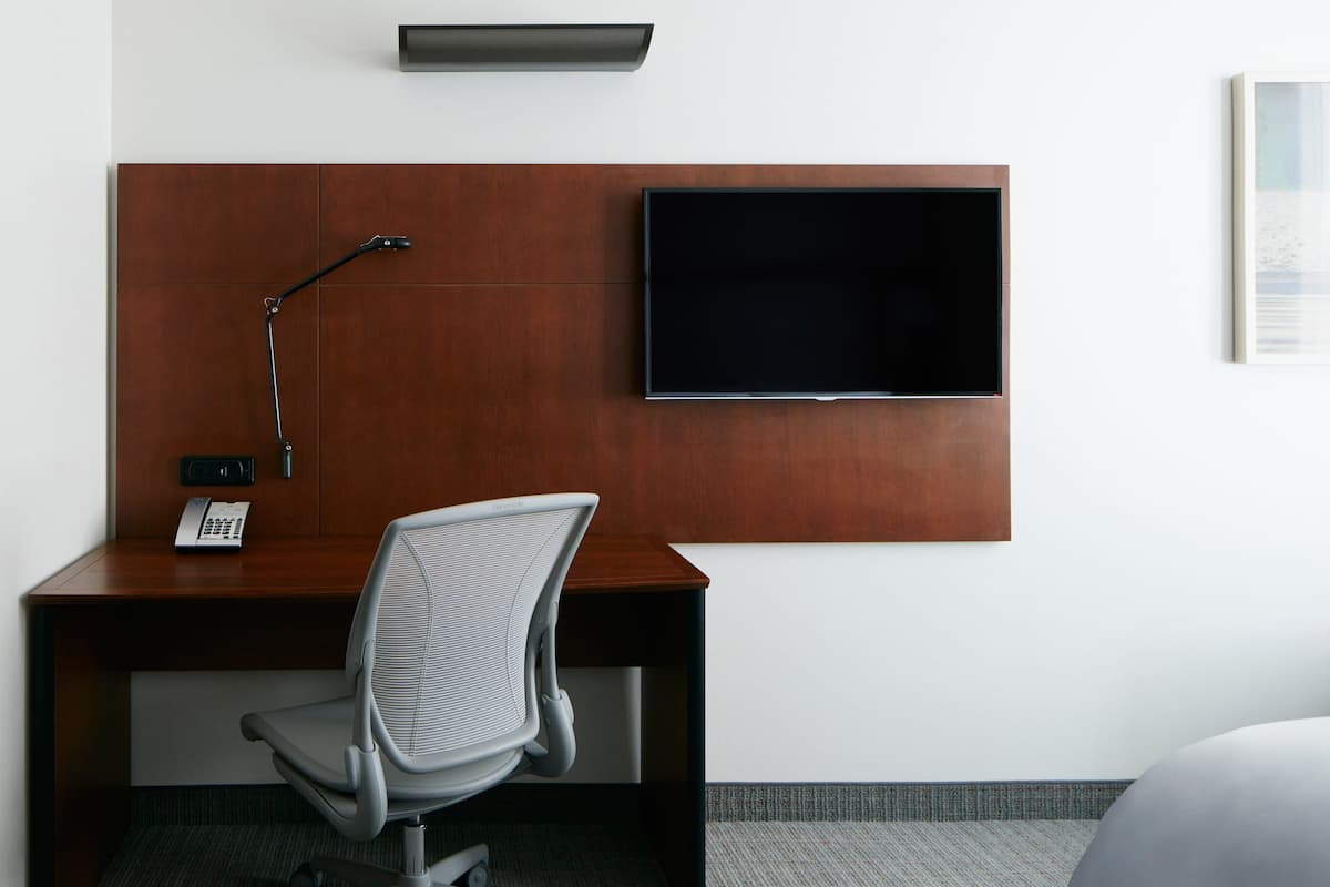 A hotel room with a wooden desk, a modern chair, a flat-screen TV mounted on the wall, and a telephone on the desk.