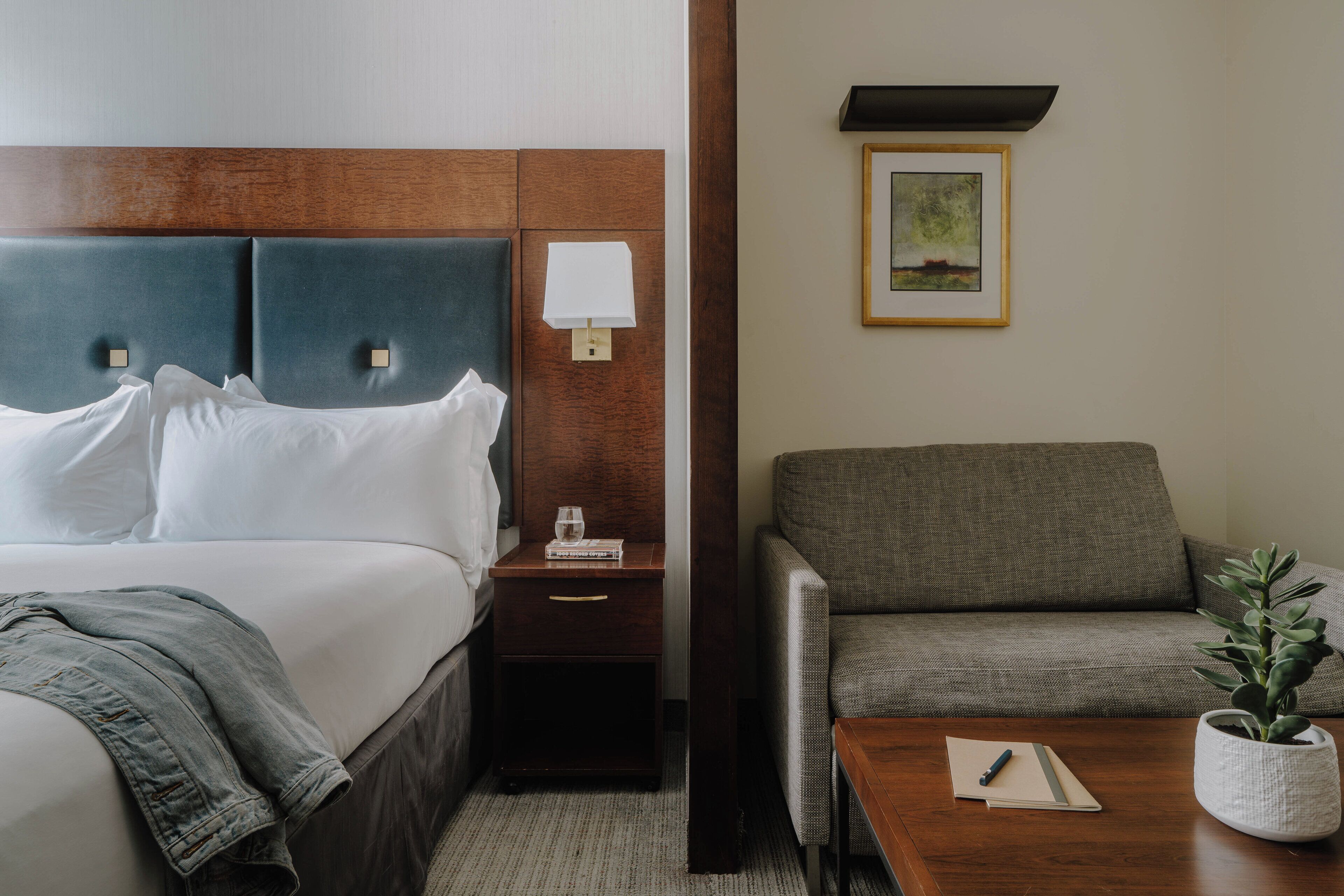 A hotel room with a bed, a nightstand, a lamp, a framed picture on the wall, a sofa, and a small table with a plant.
