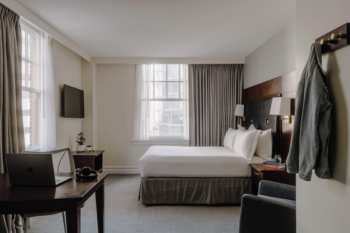 A hotel room with a bed, a desk with a laptop, a TV, and a window with curtains.