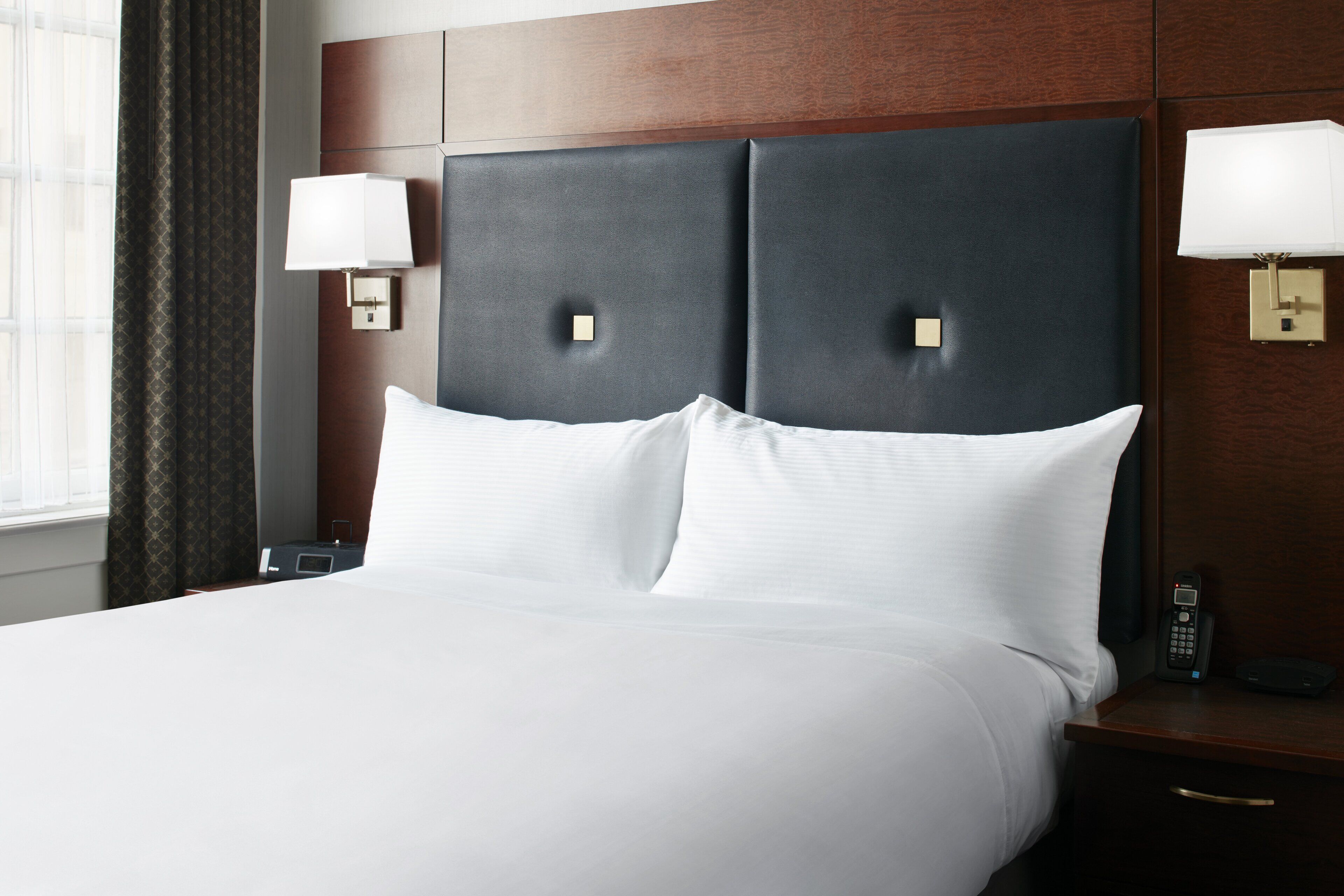 A hotel room with a large bed, two bedside lamps, a nightstand with a phone, and a window with curtains.