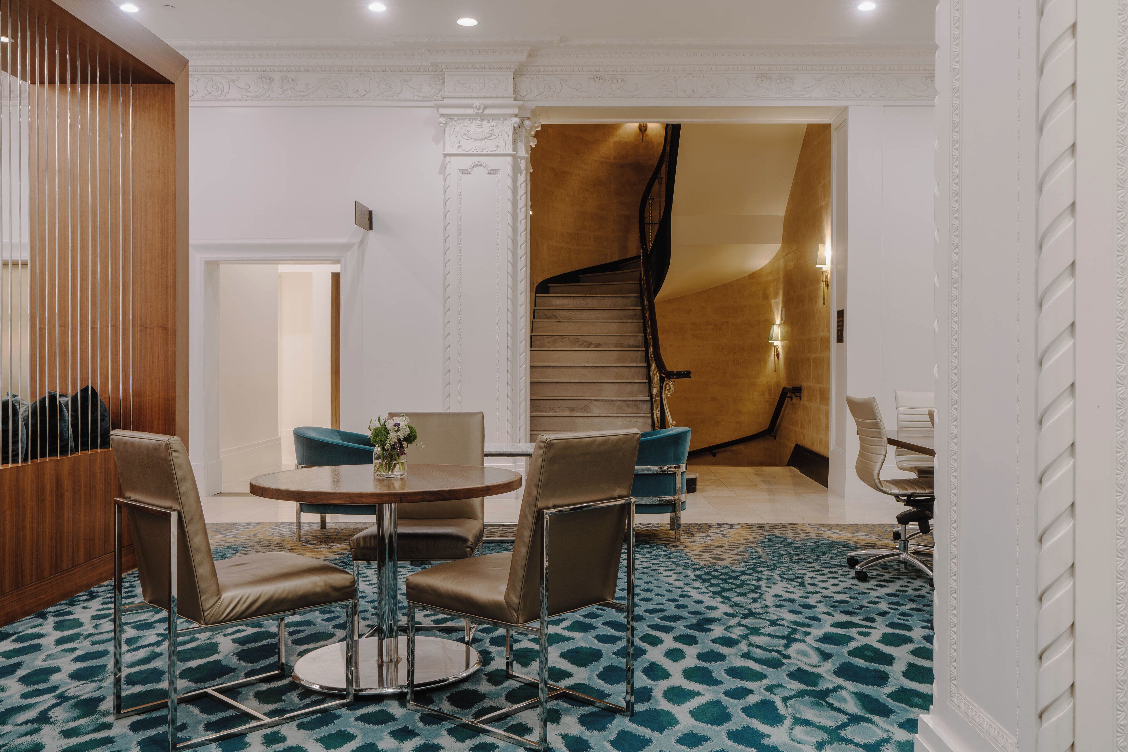 A hotel lobby with a round table, two chairs, a staircase, and a patterned carpet.