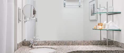 A modern bathroom with a granite countertop sink, a round mirror, a toilet, and a glass shelf with toiletries.