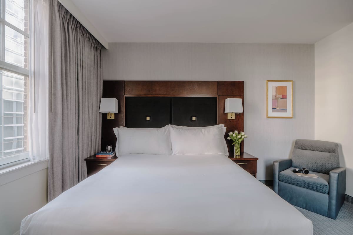 A hotel room with a large bed, two bedside tables with lamps, a chair, a framed picture on the wall, and a window with curtains.