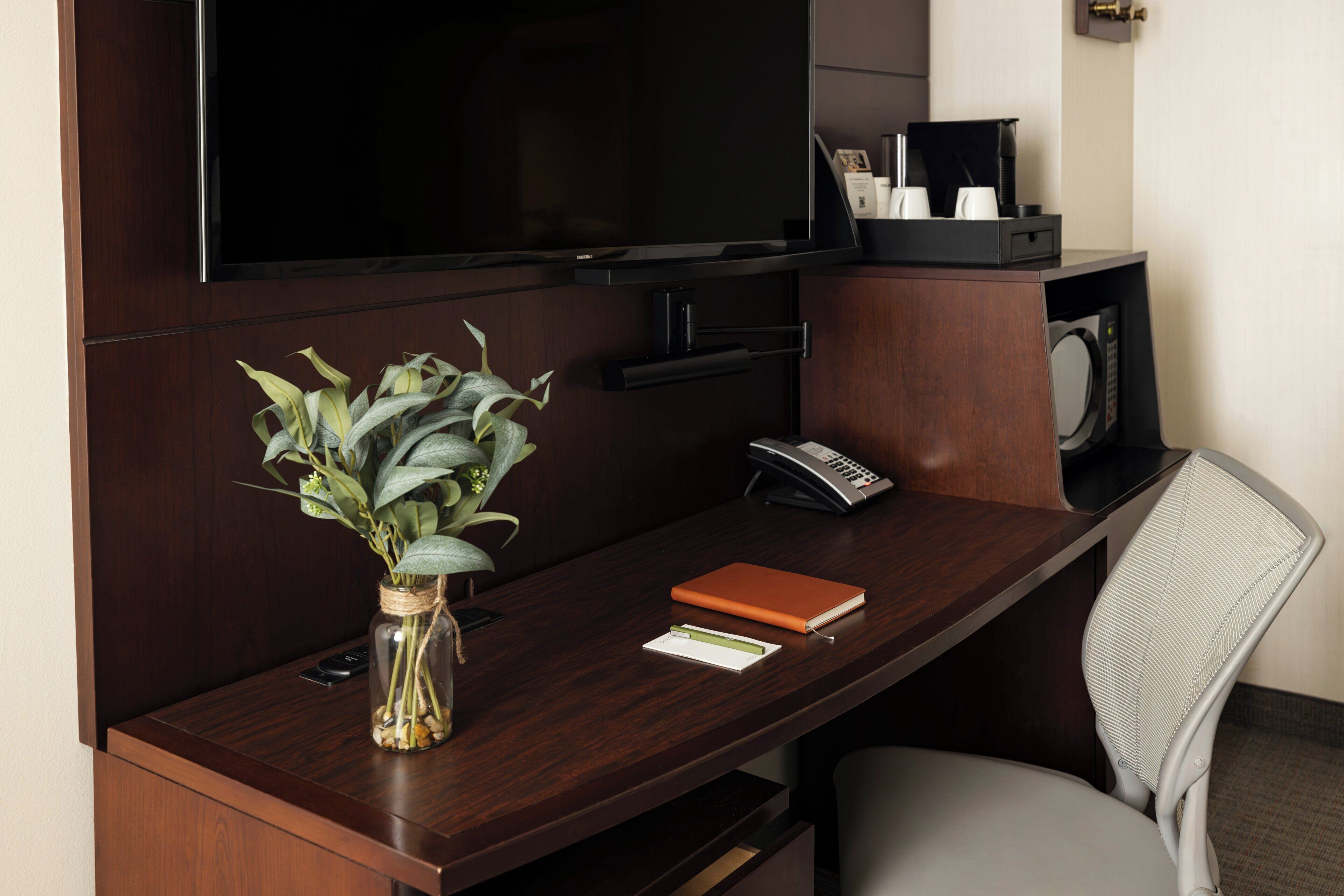 A hotel room desk with a vase of green plants, a notebook, a pen, a telephone, and a flat-screen TV mounted on the wall.