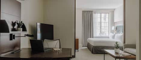A hotel room with a bed, a desk with a laptop, a TV, and a small table with a plant.