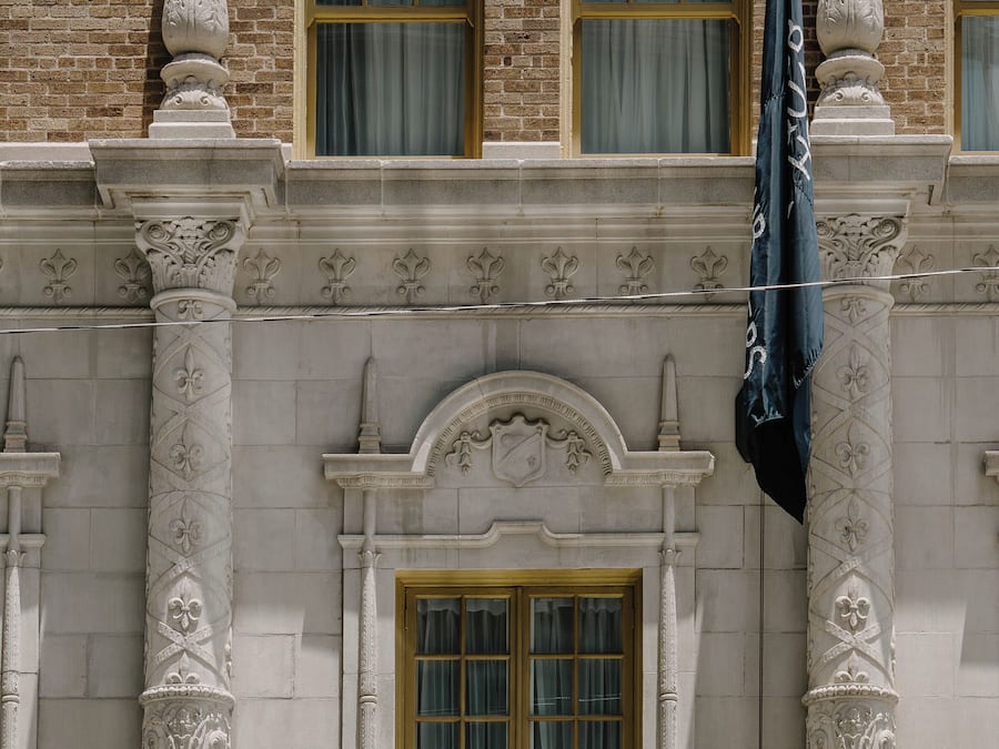 A building facade with three windows, each with a wooden frame and green curtains. Decorative columns with intricate carvings flank the windows.