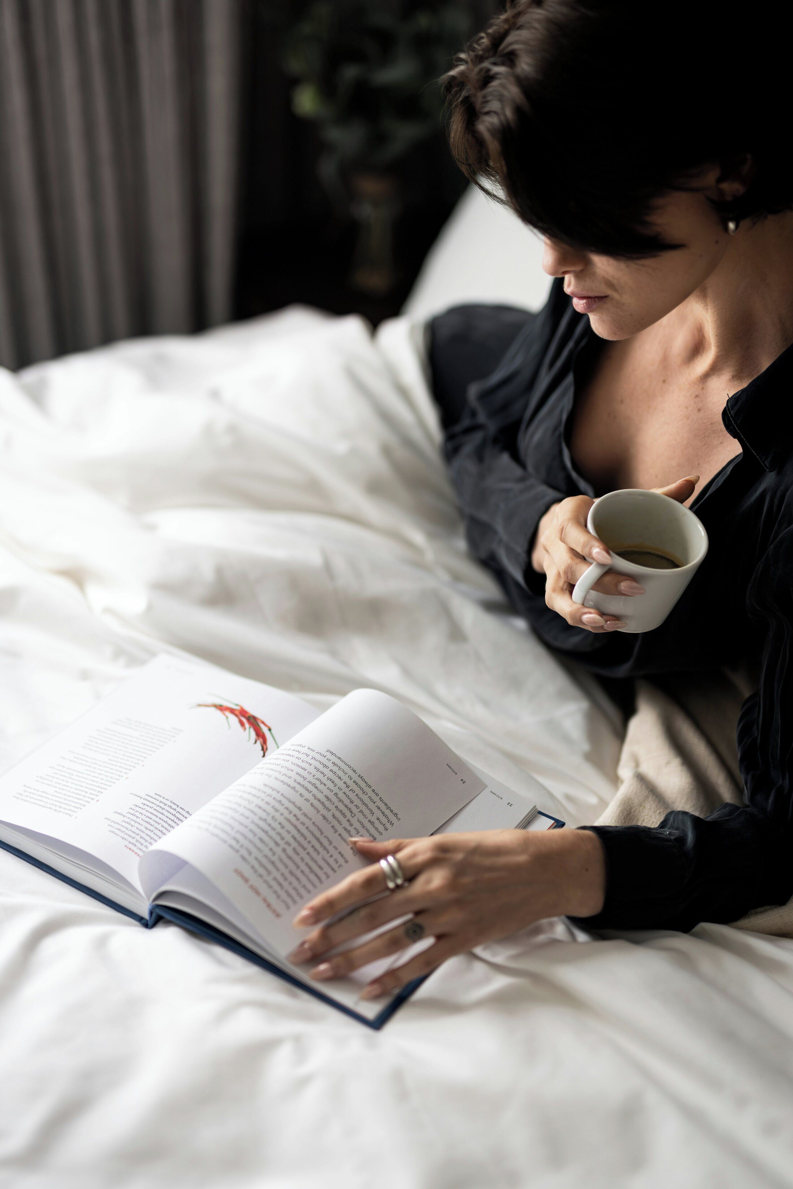 A person lying in bed reading a book with a cup of coffee.
