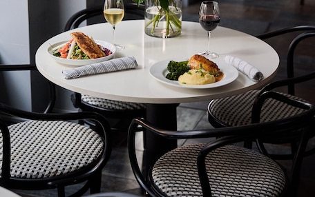 A dining table with white tablecloth, two plates of food, two wine glasses, and a vase with white flowers.