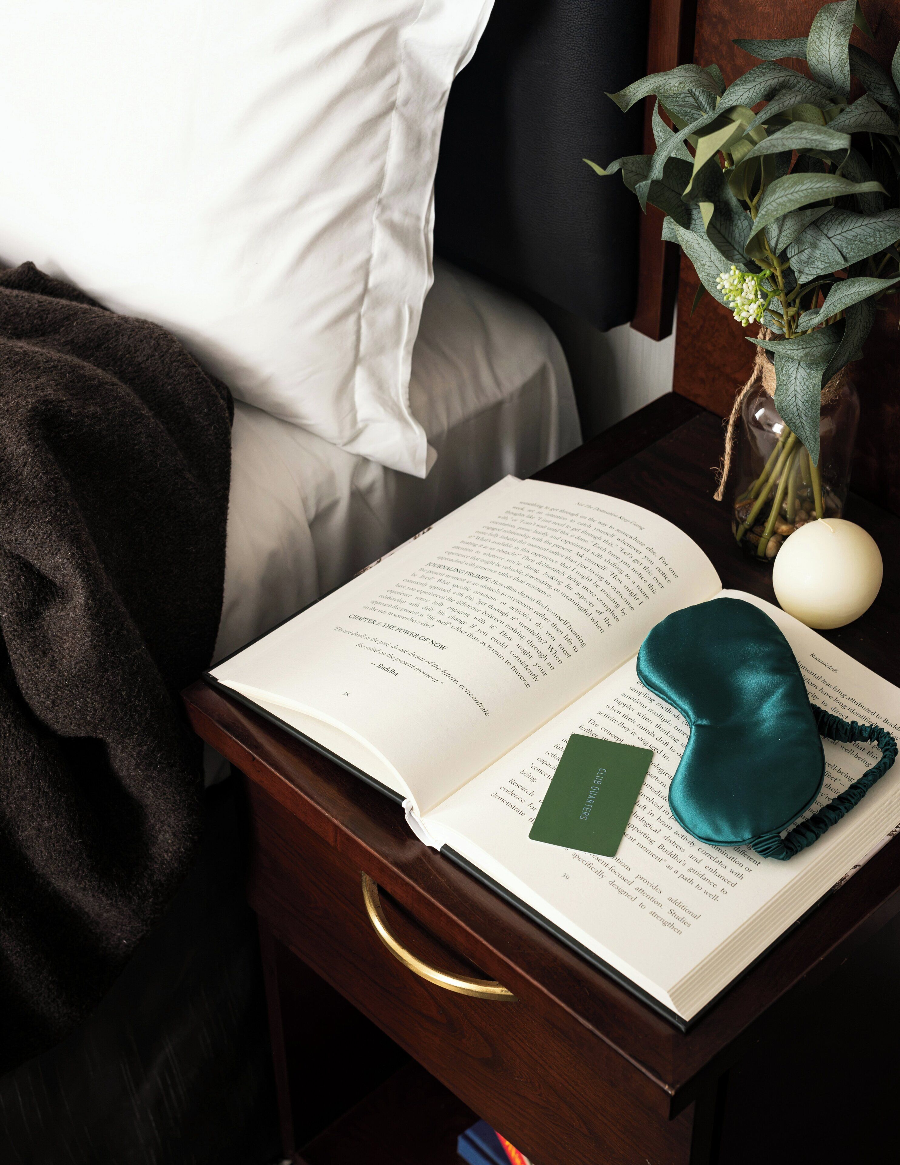 Un livre ouvert sur une table de chevet en bois, avec une carte verte, un coussin turquoise et un vase contenant des feuilles vertes et des fleurs blanches.