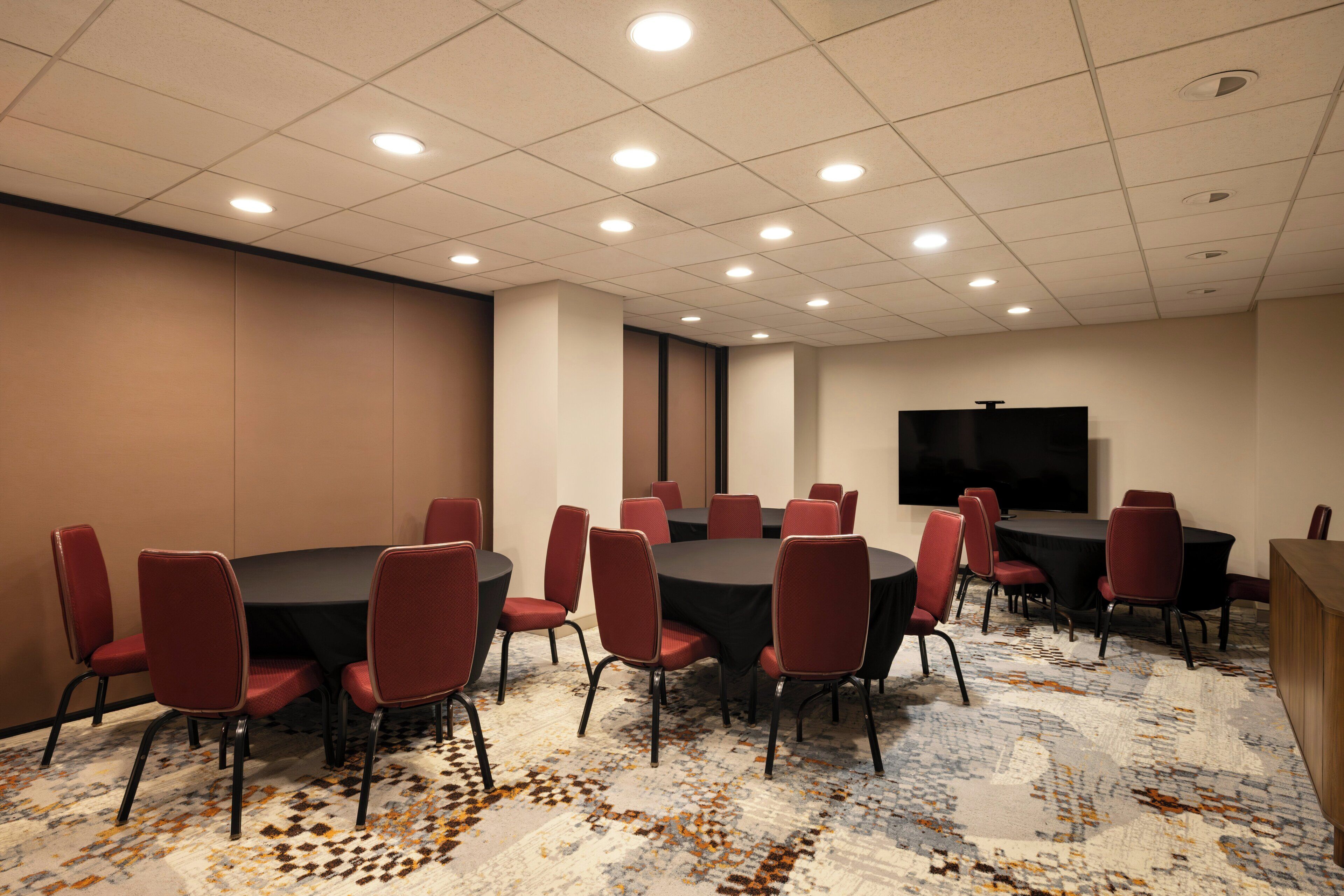A conference room with round tables and red chairs, a flat-screen TV on the wall, and patterned carpet.