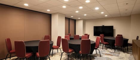 A conference room with round tables and red chairs, a flat-screen TV on the wall, and patterned carpet.