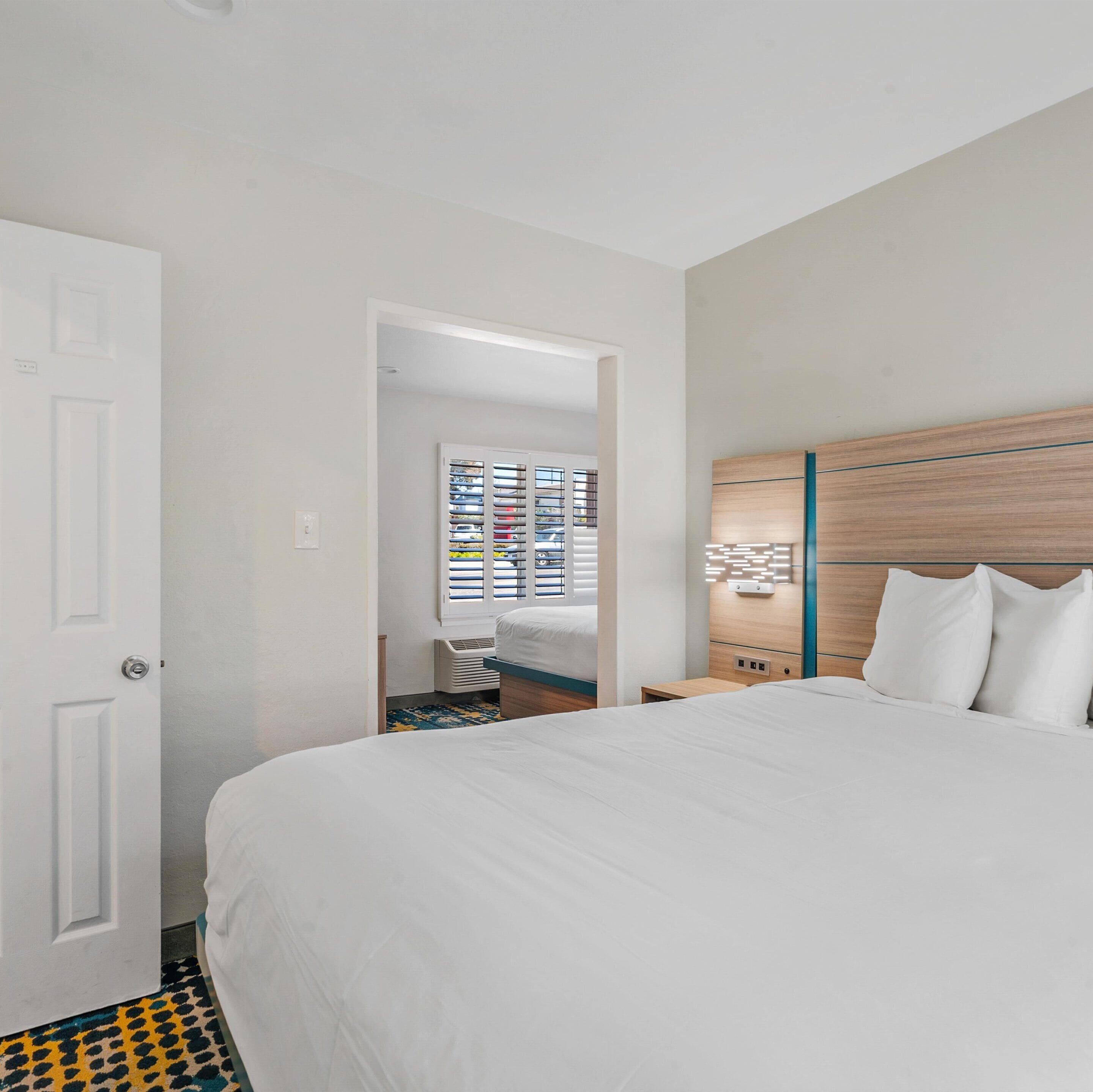 A hotel room with a large bed, a wooden headboard, a nightstand, and a view of a cityscape through the window.