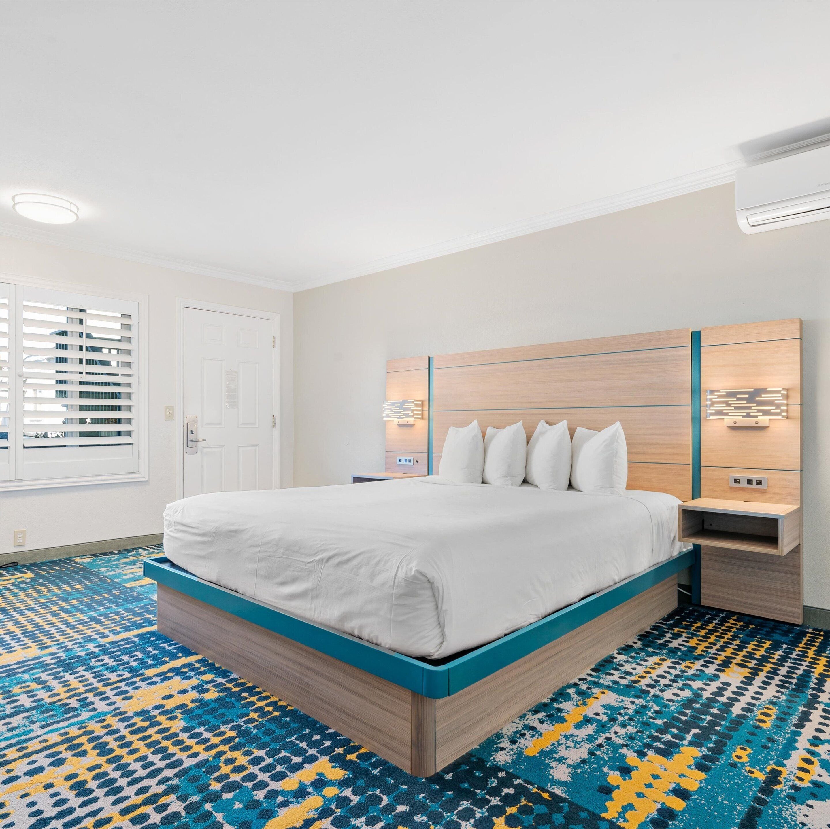 Habitación de hotel moderna con una cama grande, cabecera de madera, alfombra estampada y una ventana con persianas.