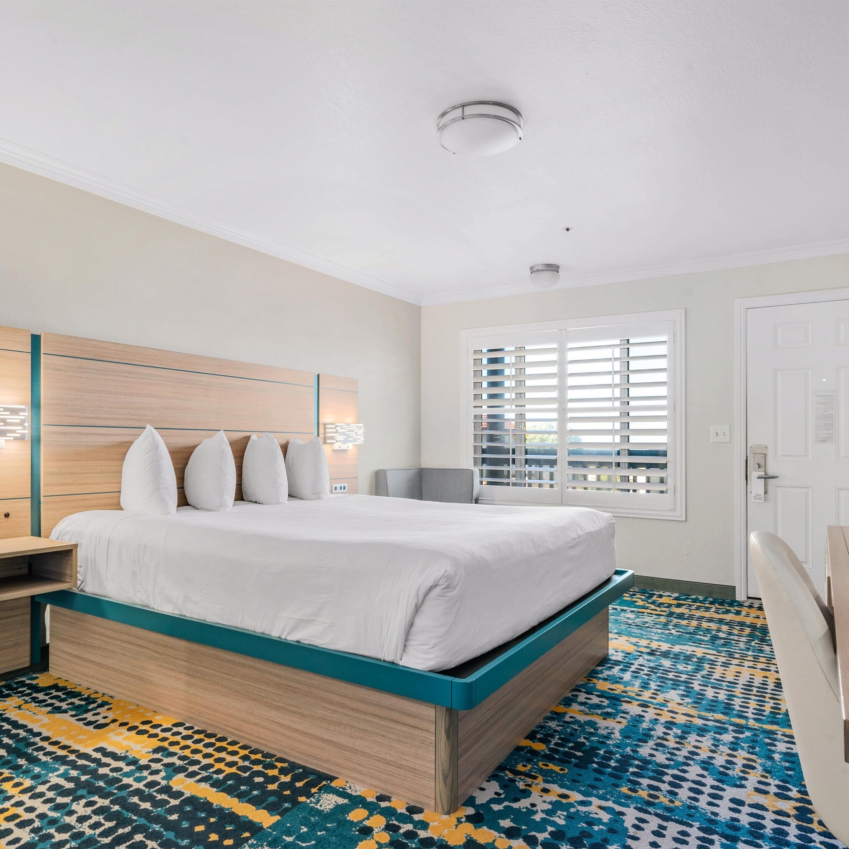 A hotel room with a bed, bedside tables, a window with blinds, and a patterned carpet.