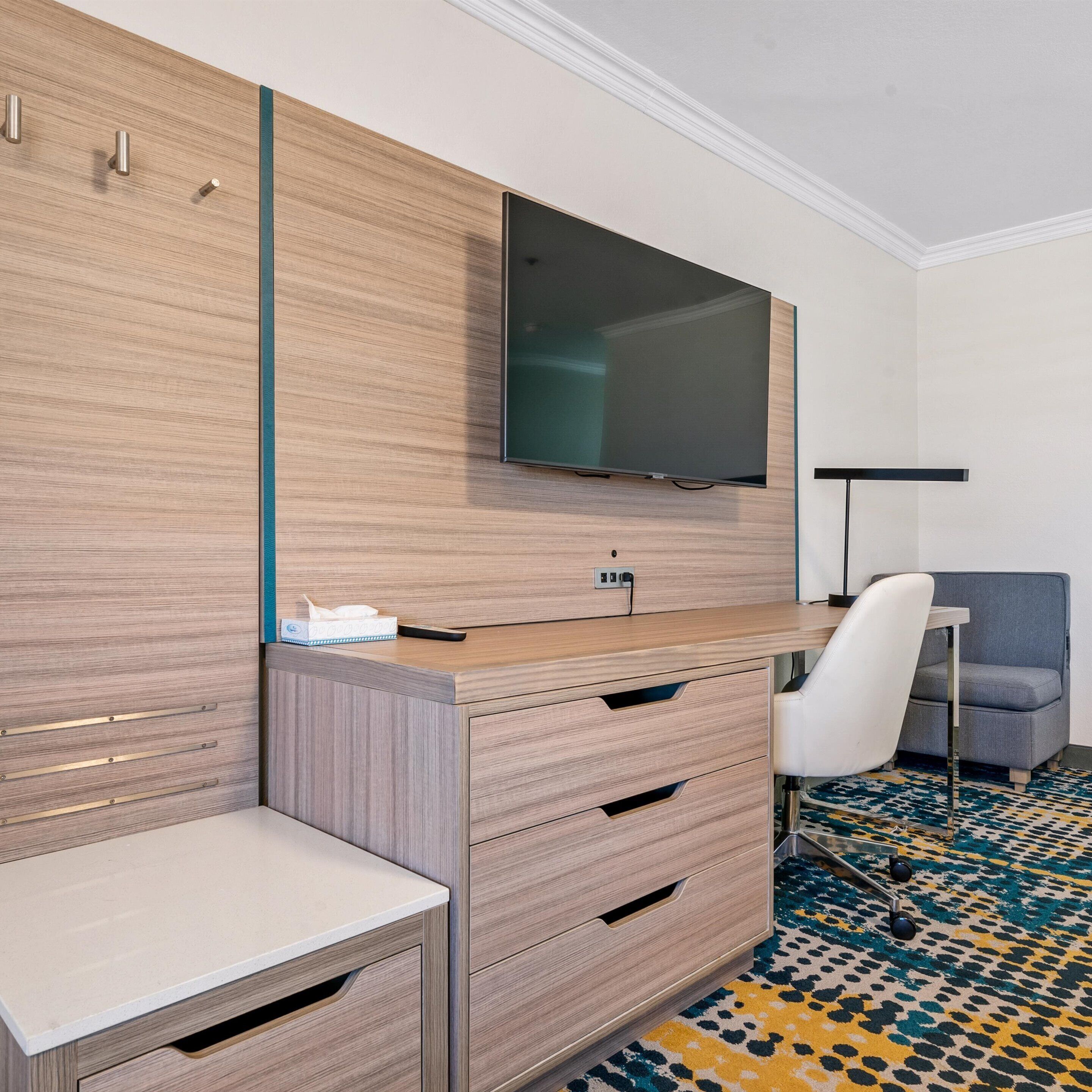 Une chambre d’hôtel moderne avec un bureau en bois, un téléviseur à écran plat fixé au-dessus, un fauteuil gris et une moquette à motifs.