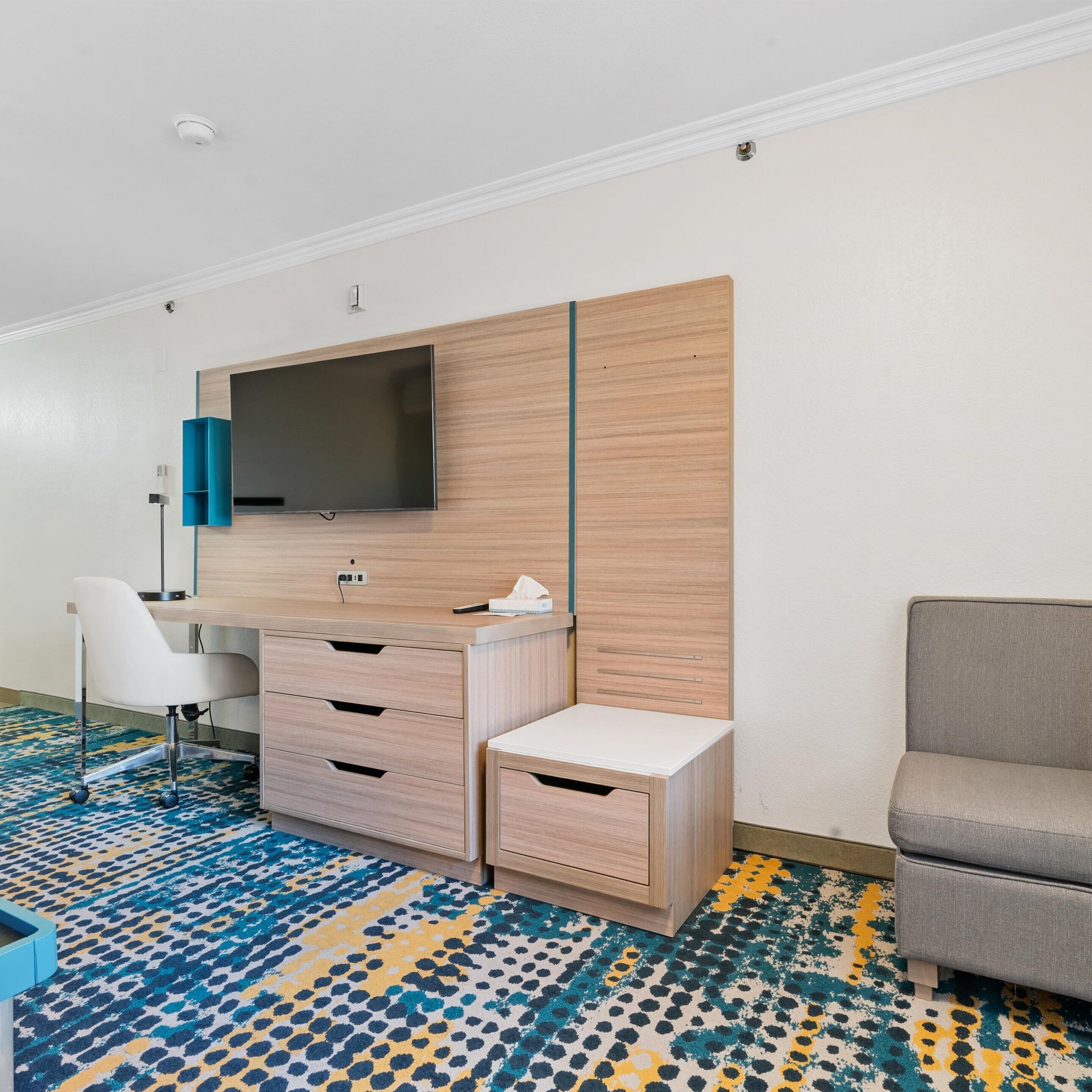 A modern hotel room with a flat-screen TV mounted on a wooden panel wall, a white desk with drawers, a grey sofa, and a patterned carpet.