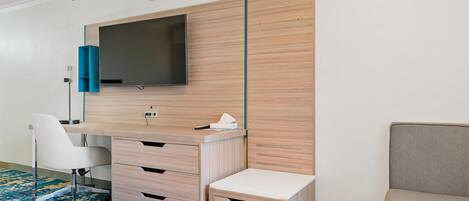 A modern hotel room with a flat-screen TV mounted on a wooden panel wall, a white desk with drawers, a grey sofa, and a patterned carpet.
