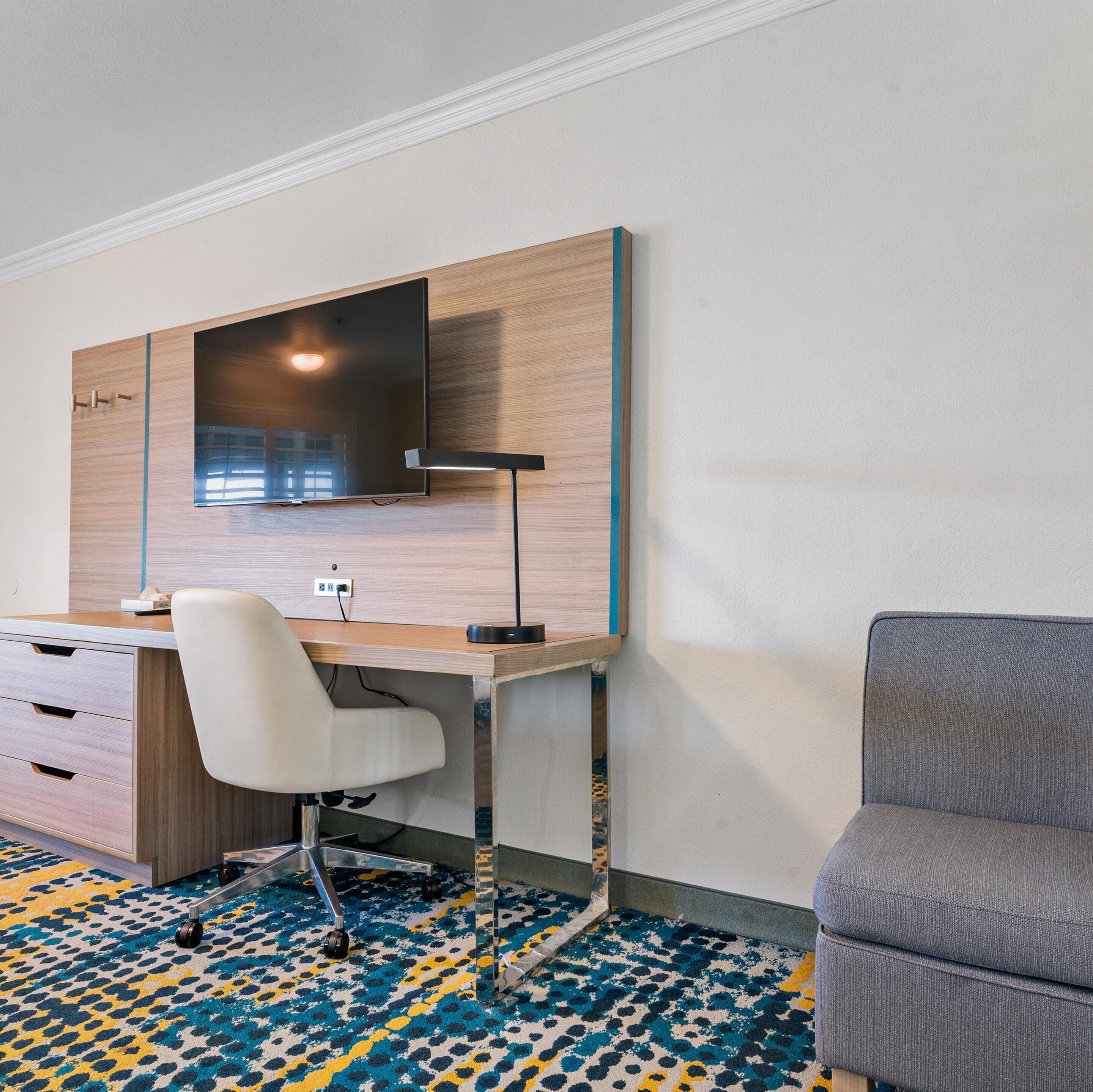 Una habitación de hotel moderna con un escritorio de madera, un televisor de pantalla plana, una silla blanca y un sofá gris.