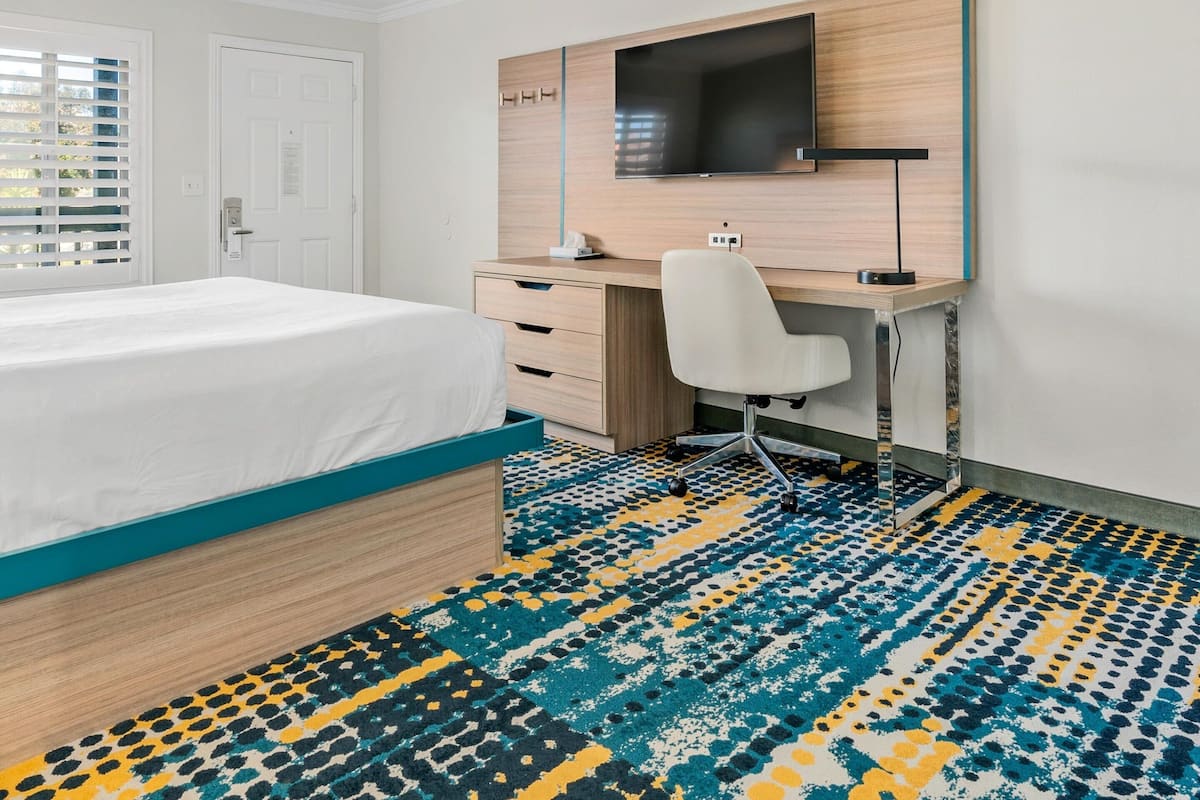A hotel room with a bed, a desk with a chair, a TV mounted on the wall, and a patterned carpet.