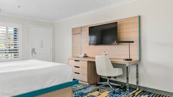 A hotel room with a bed, a desk with a chair, a TV mounted on the wall, and a patterned carpet.