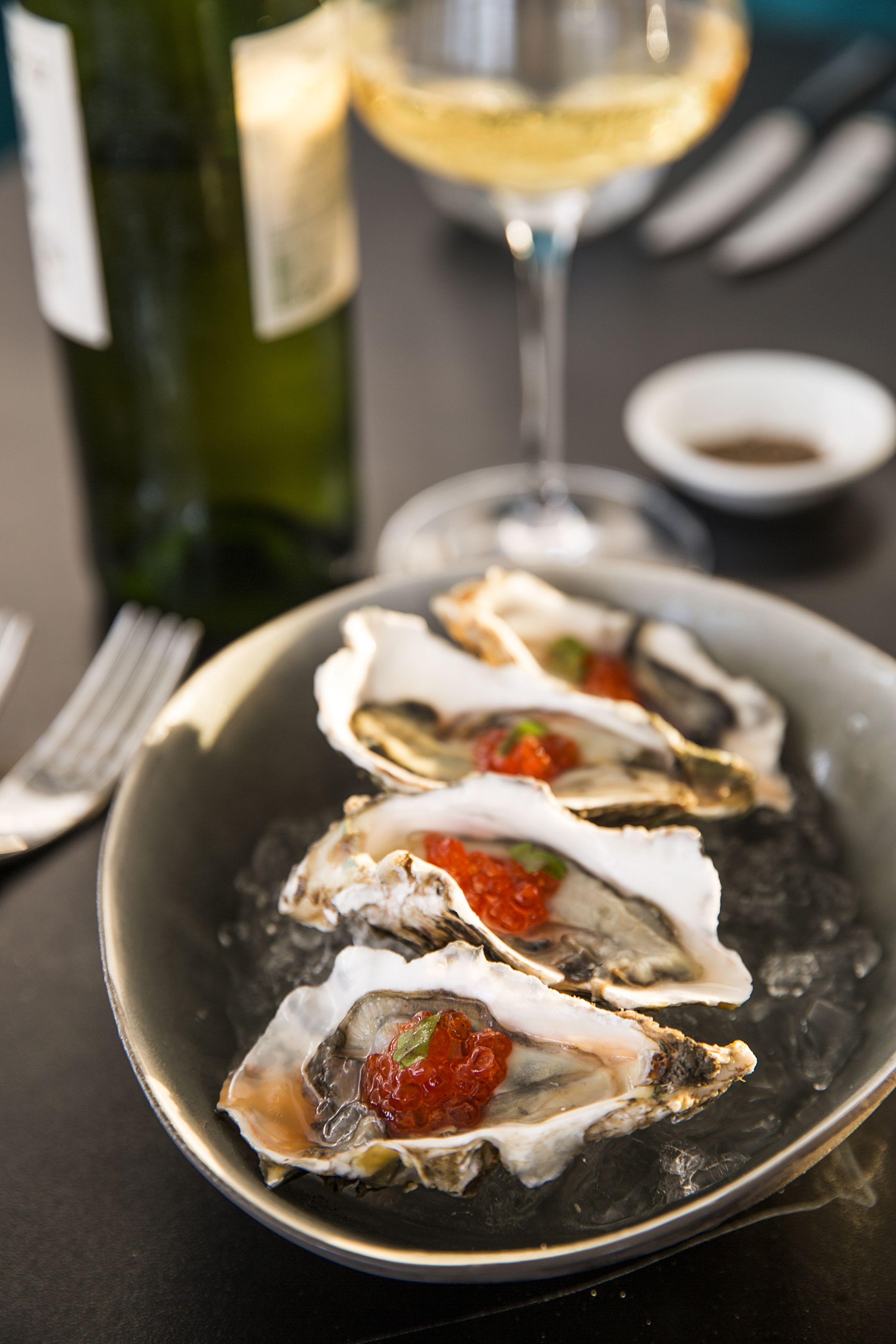 A plate of oysters with a red topping, a glass of white wine, and a bottle of wine on a table.