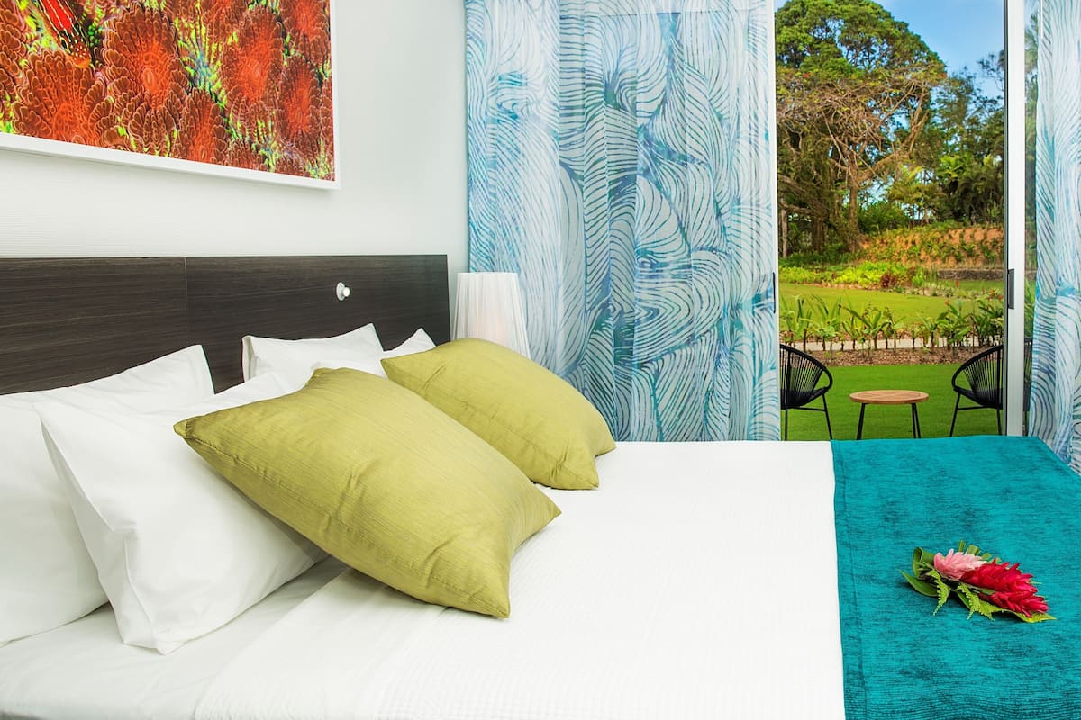 Een hotelkamer met een bed, twee kussens, een blauwgroene sprei, een stoel en uitzicht op een tuin door gordijnen.